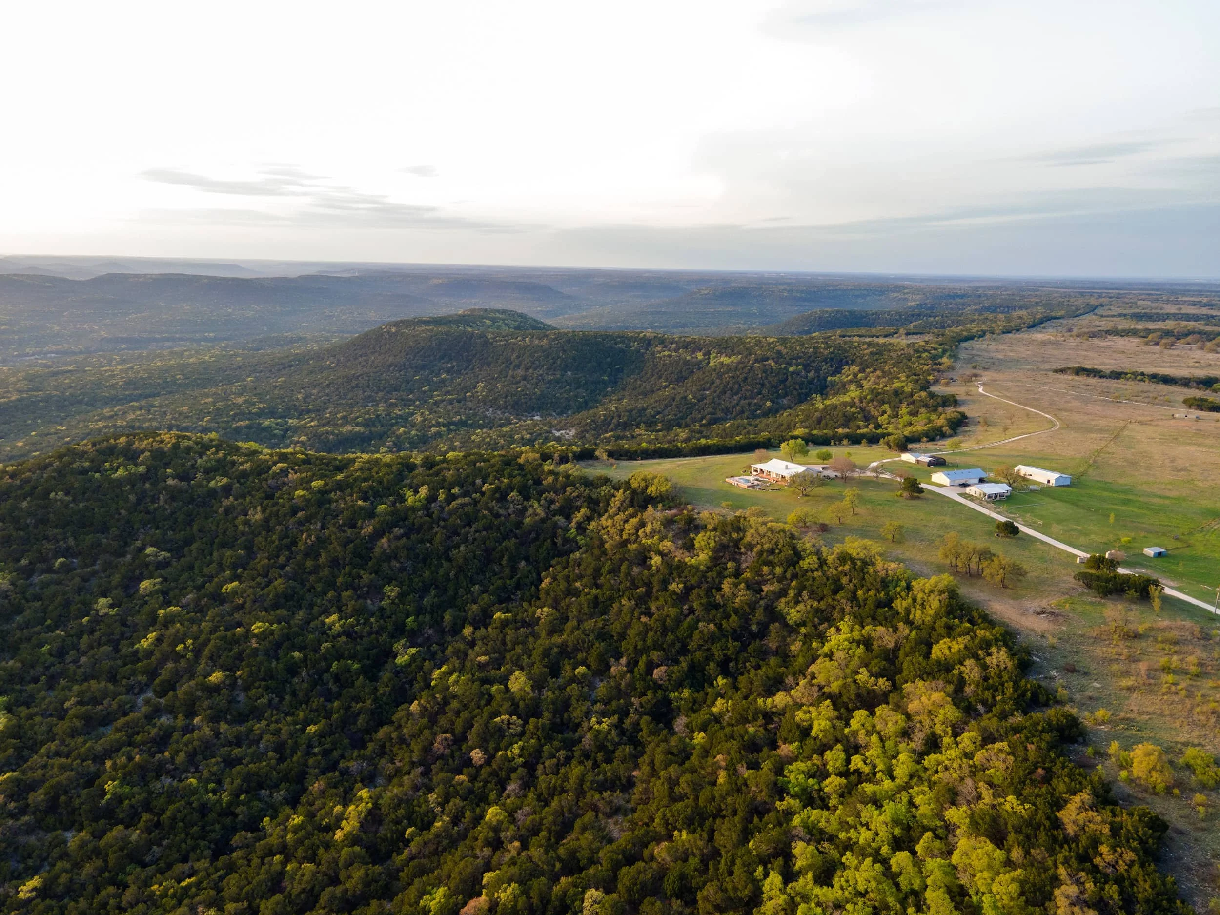 Aerial view of expansive green forested hills, open fields, and a few farm buildings under a cloudy sky.
