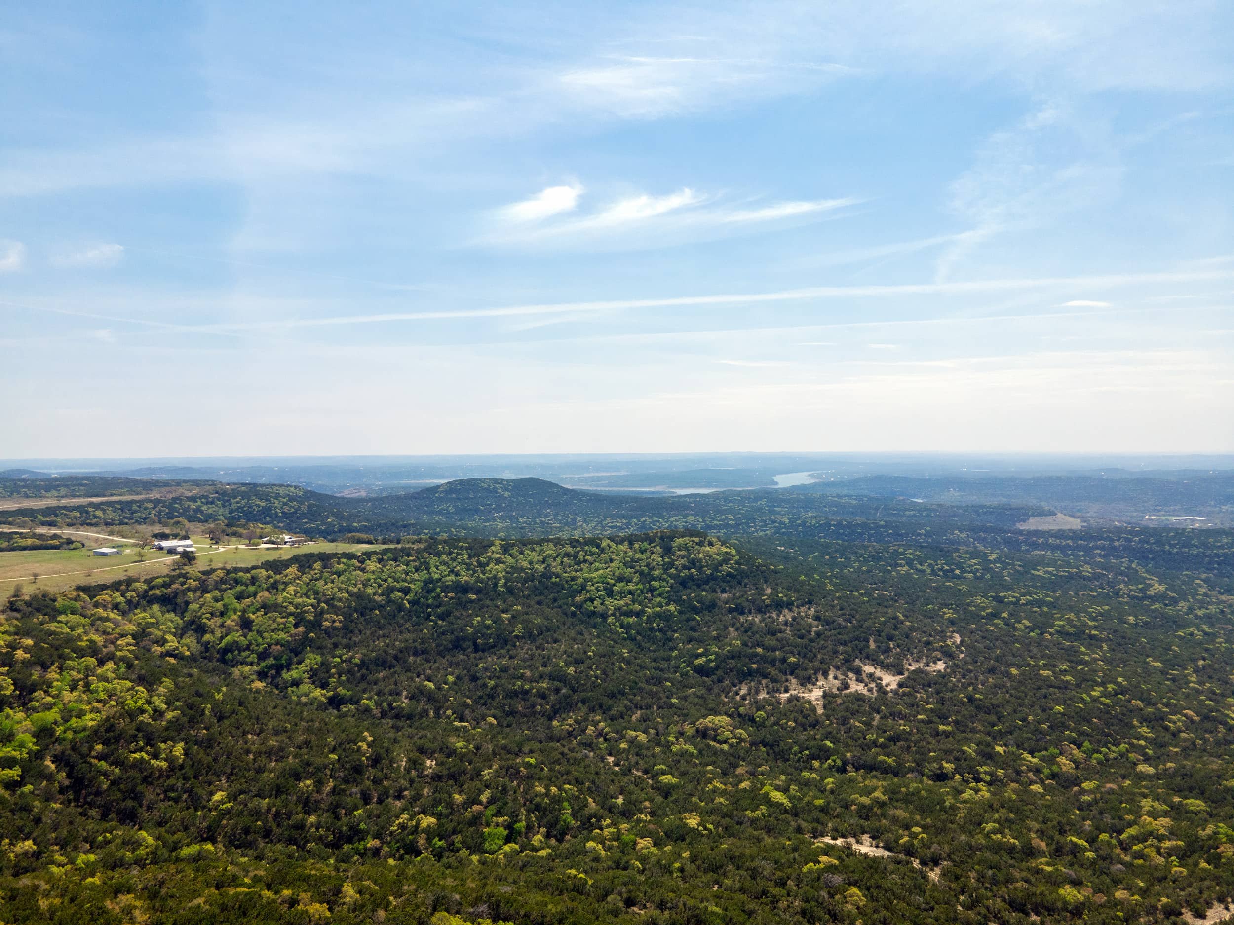 A broad view of a lush green landscape with forests and open fields, extending to a distant body of water under a partly cloudy sky.