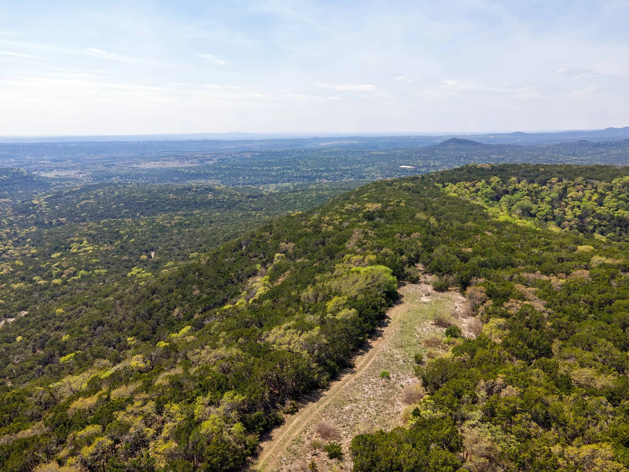 Aerial view of a lush green forested landscape with a dirt trail running through it, rolling hills in the distance, and a partly cloudy sky overhead.