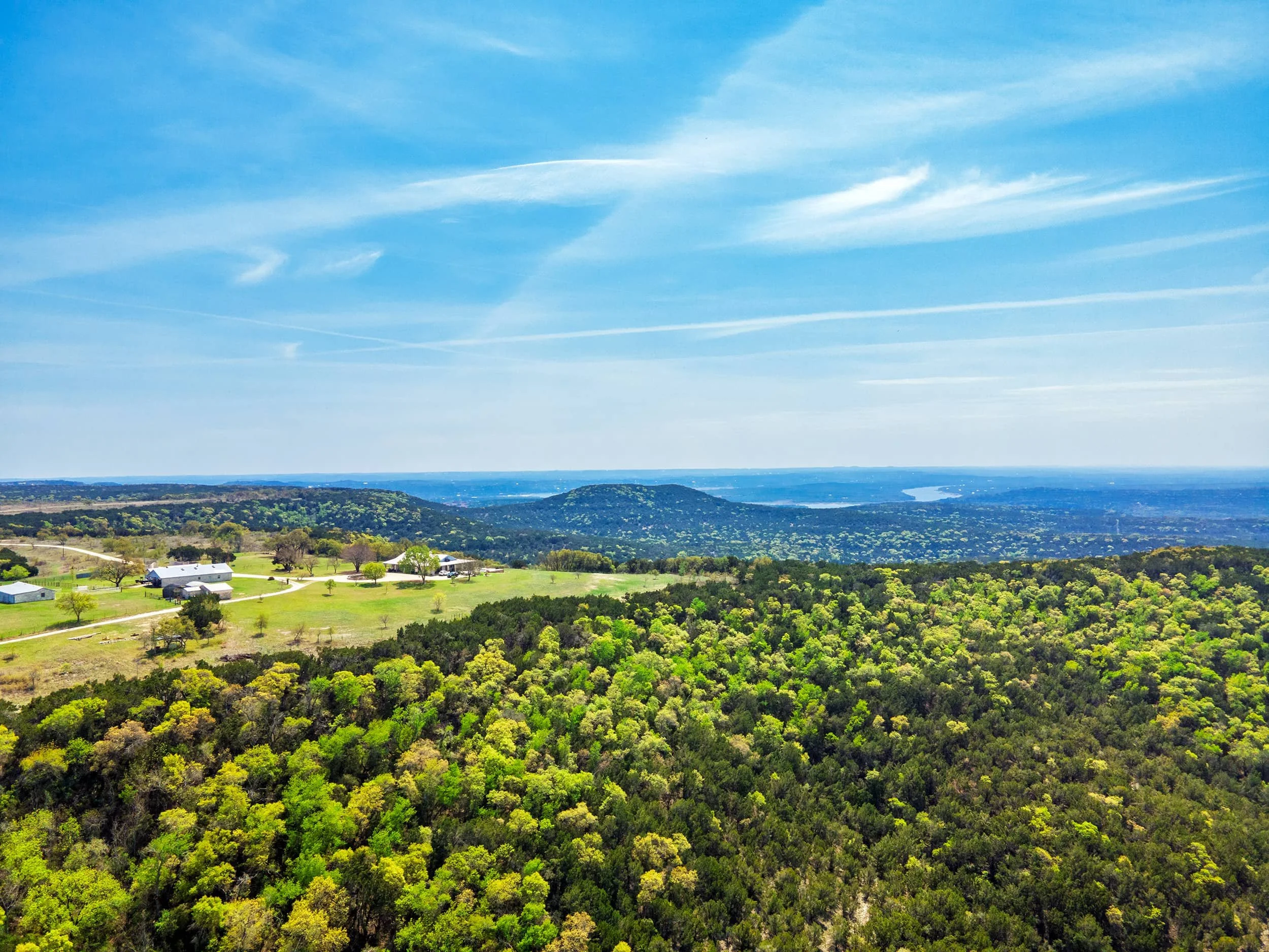 A landscape view showing a large green forest with trees on rolling hills, a few farm buildings, and a patch of blue sky with wispy clouds, extending to a distant river or lake.
