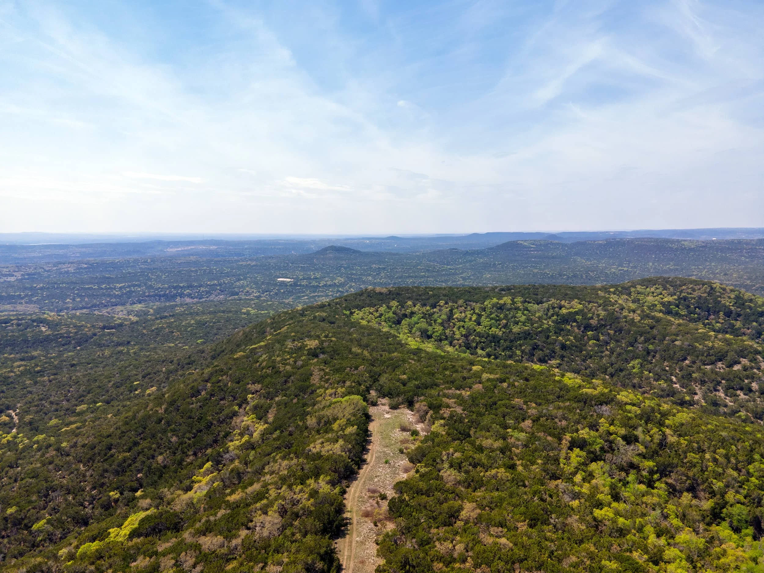 A scenic view of a wide, lush green forested landscape with a dirt trail running through it, under a partly cloudy sky.