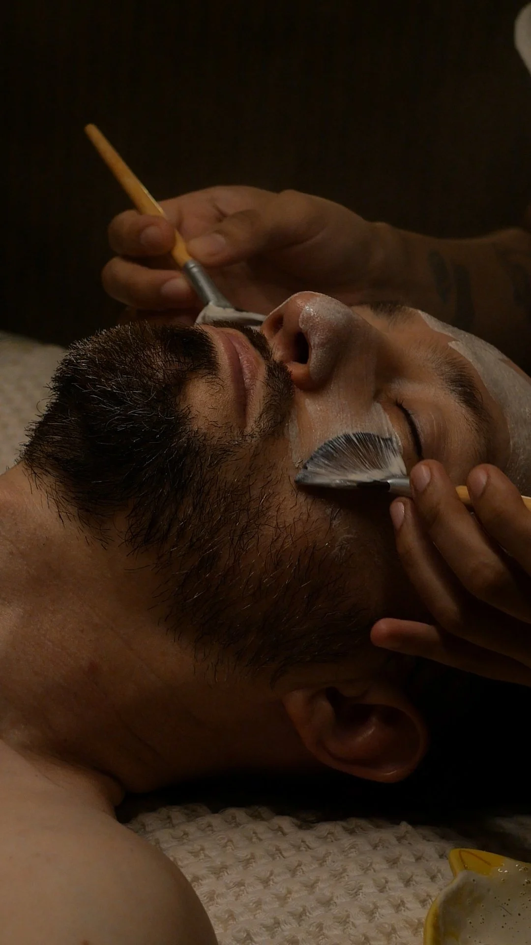 Male facial client with an ezyme exfolaition mask being applied with two brushes by esthetician.