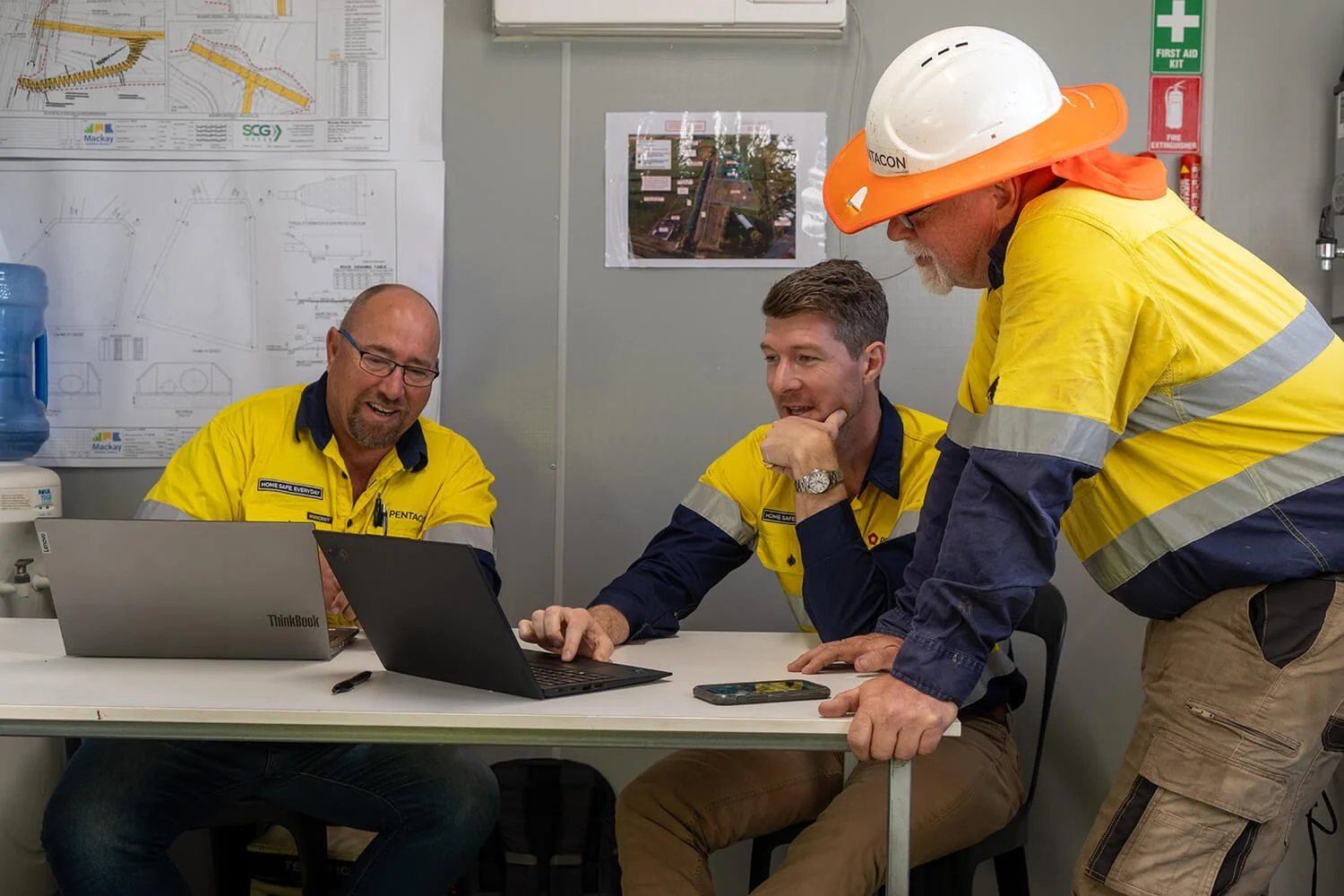 Two laptops on table with two workers in high vis clothing looking and working. One worker in high vis standing next to desk with safety hat