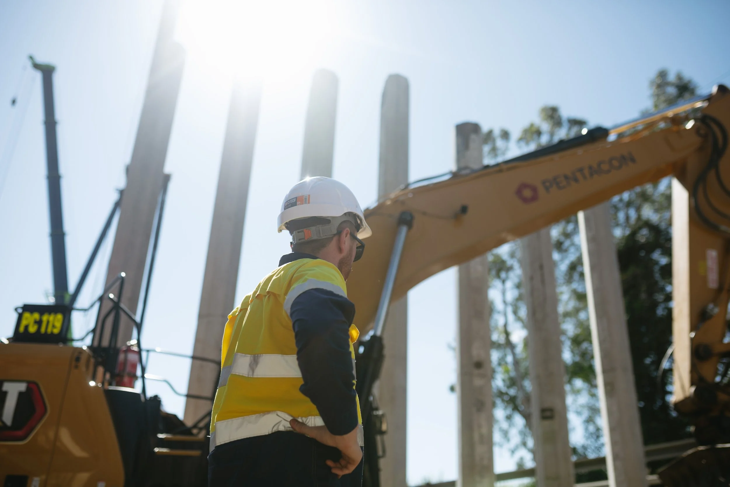 Work person in high vis and safety hard hat facing away from camera. Large machinery in background with tall concrete structure being built