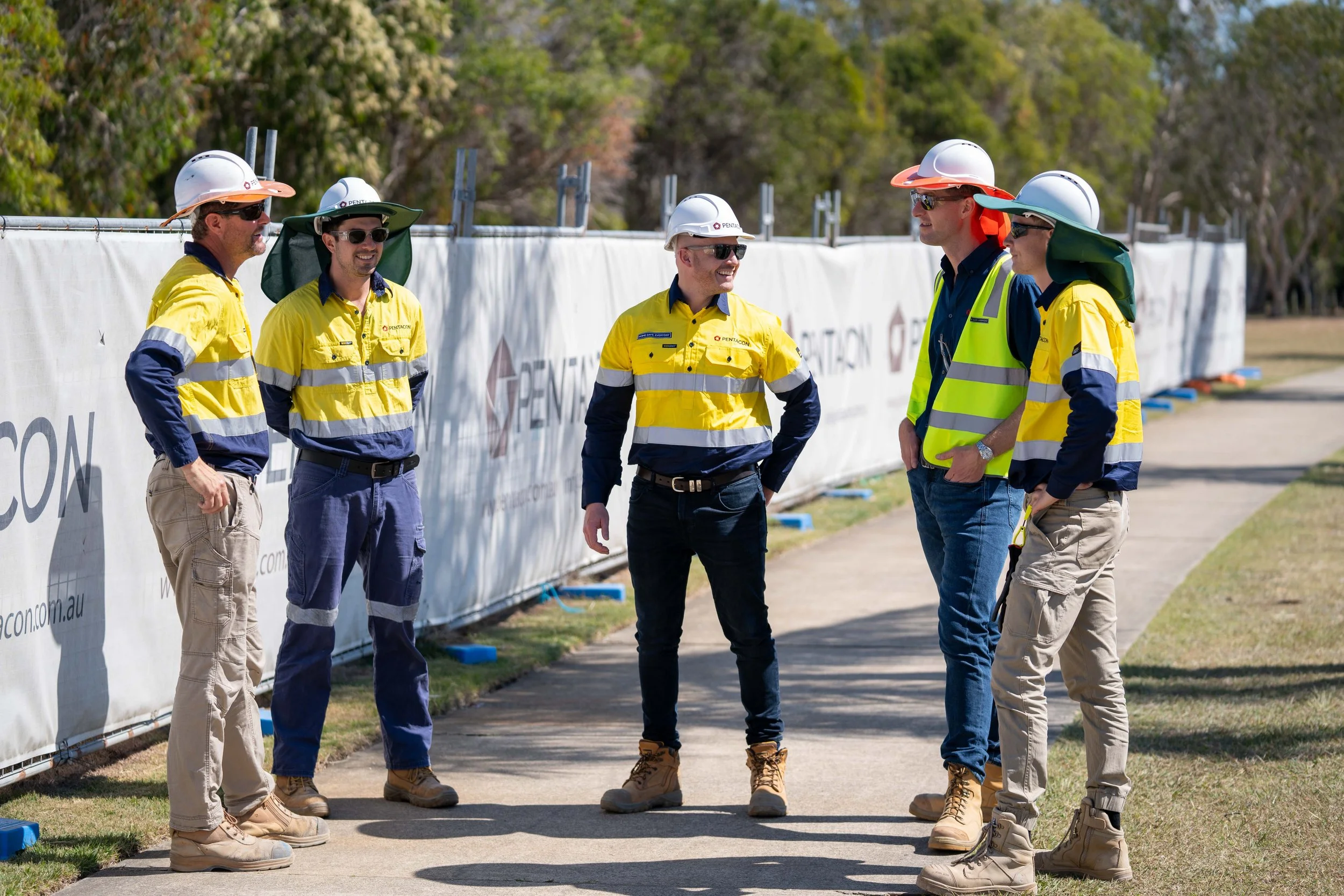 5 work personal standing in front of branded construction fence, wearing high vis and safety hard hats and sunglasses, talking to each other