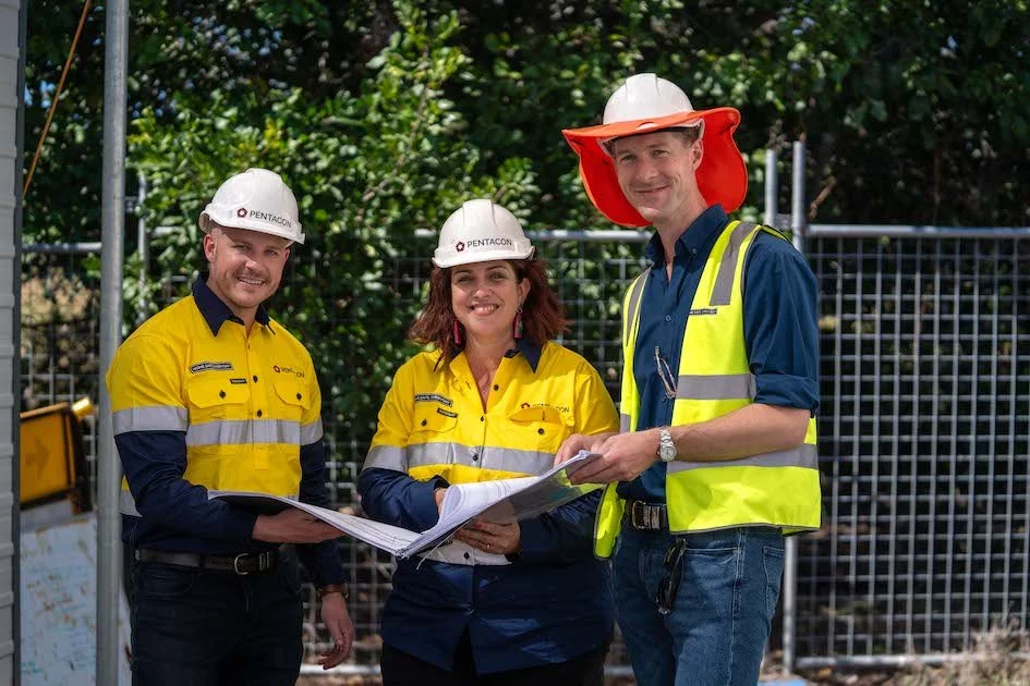 Three work person looking and smiling at camera wearing high vis and safety hard hats while holding large paperwork plans