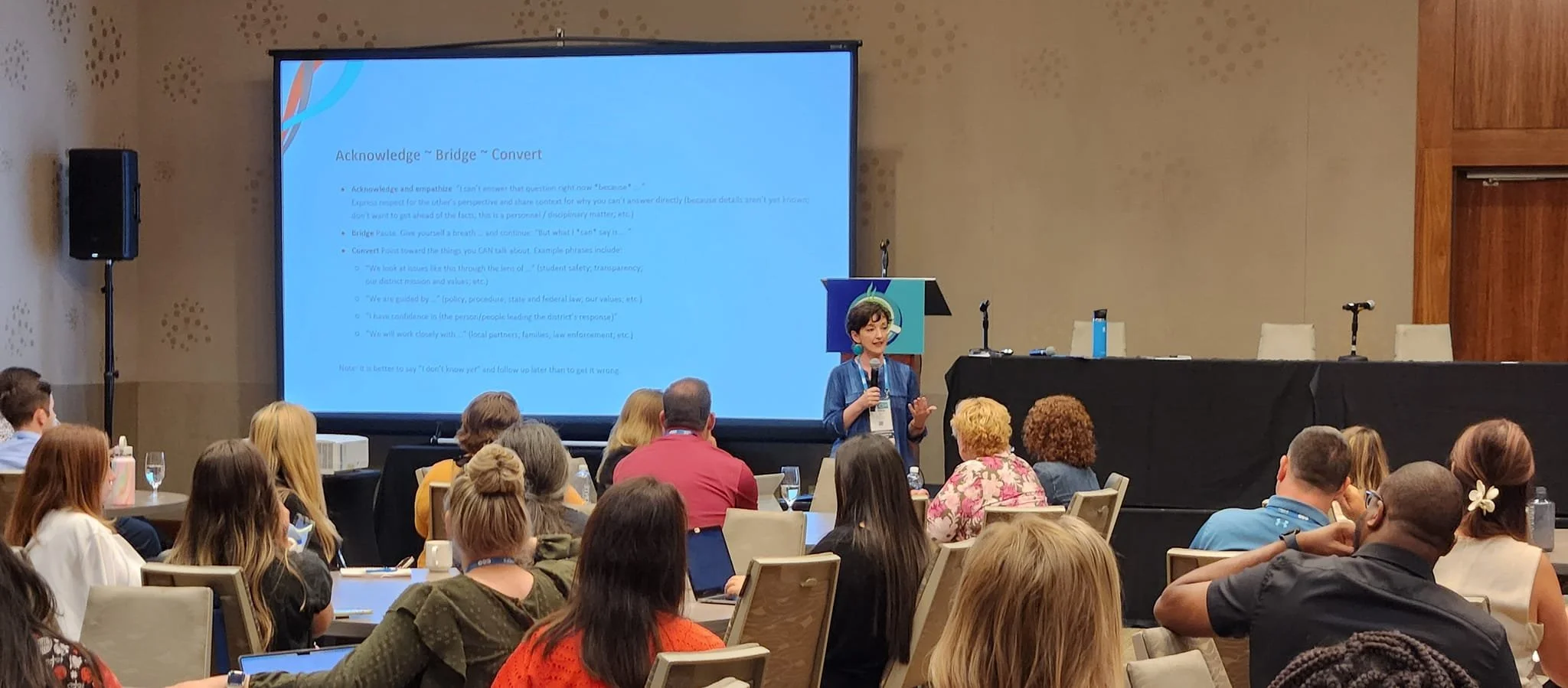 A woman giving a presentation in a conference room filled with attendees, a large screen behind her displays text and slide titles.