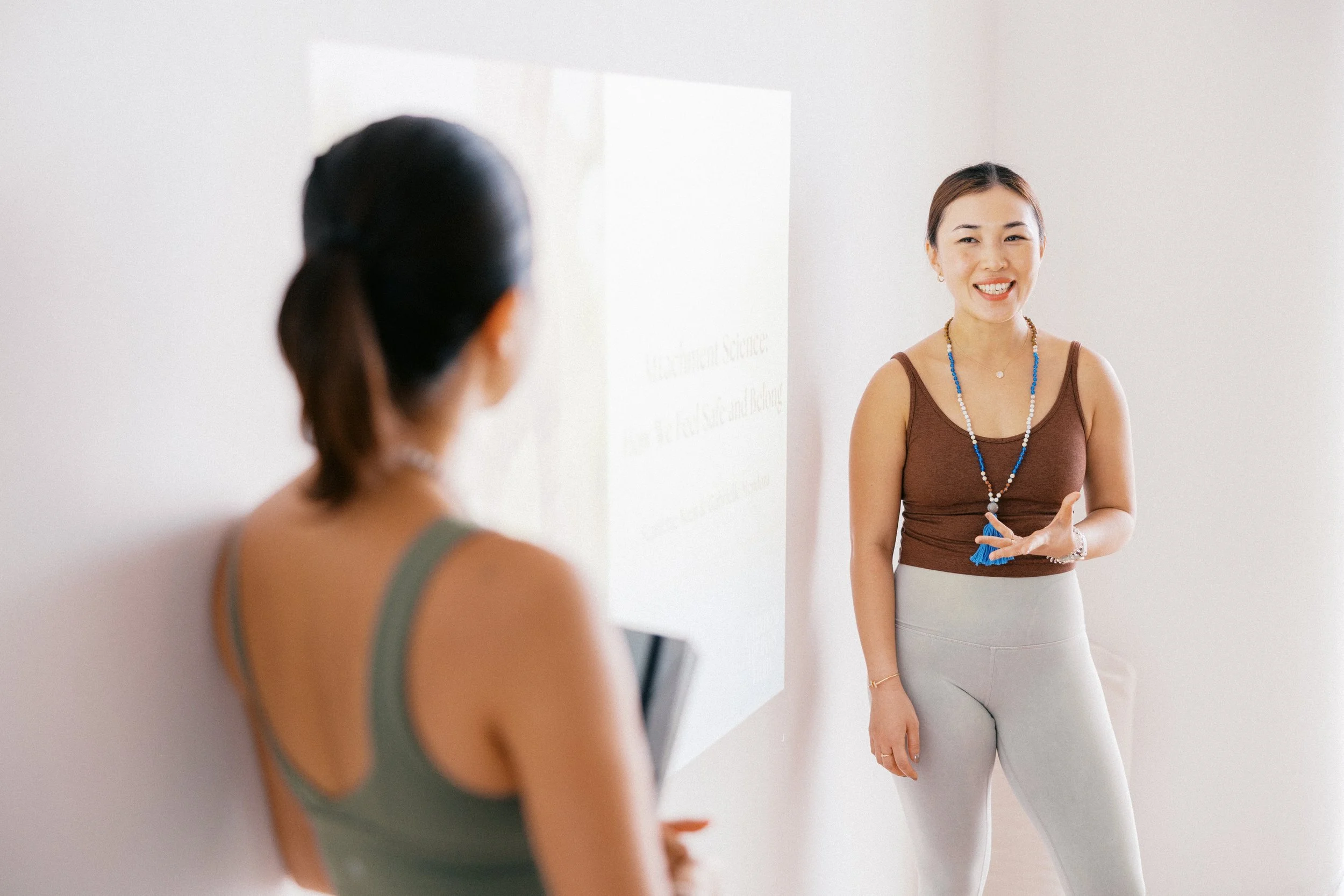 A woman with medium-length gray hair and a tan top talking to another person with their back to the camera, in a bright room with a whiteboard.