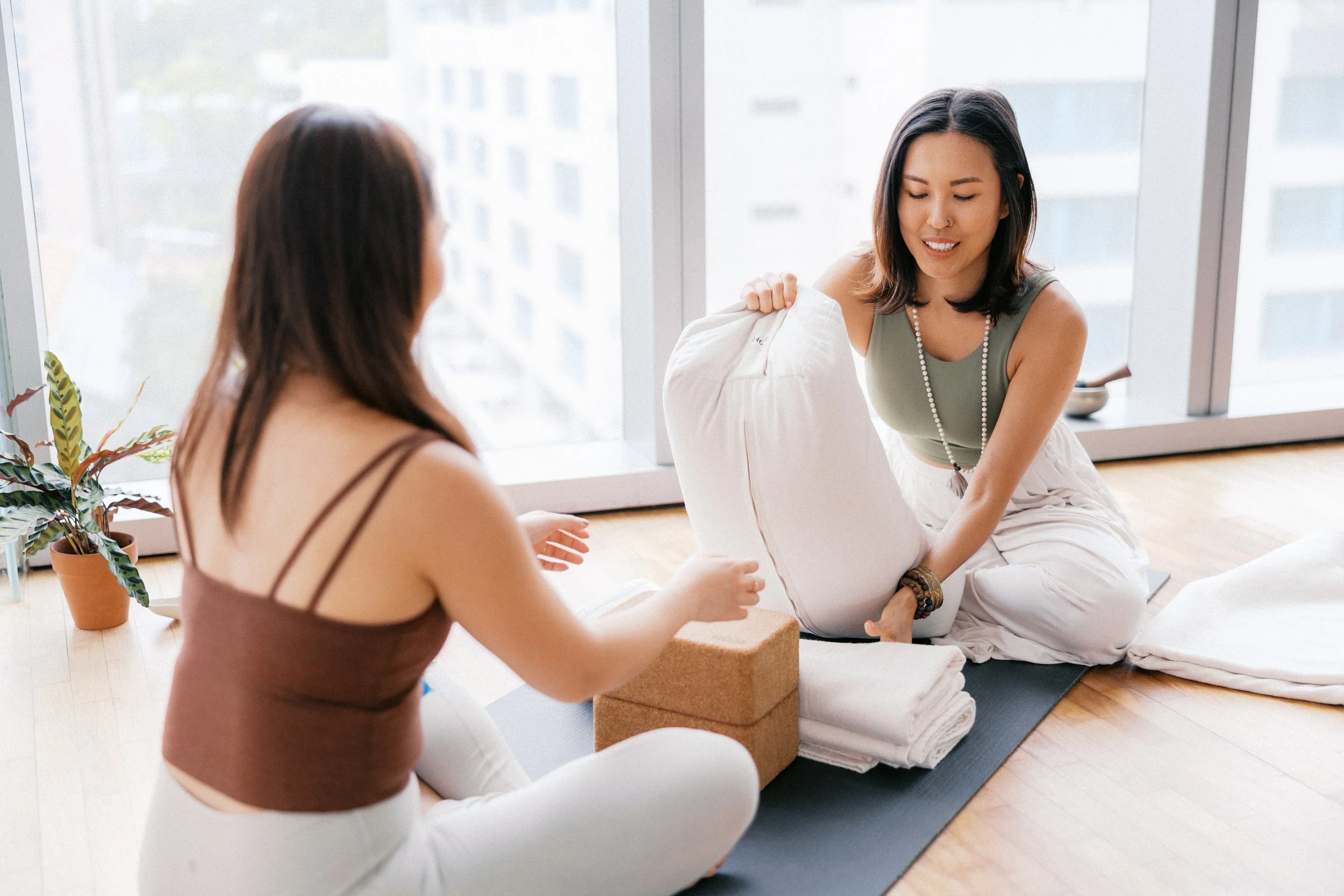 Two women sitting cross-legged on yoga mats, one is handing a rolled-up pillow to the other, with a large window and potted plant in the background.