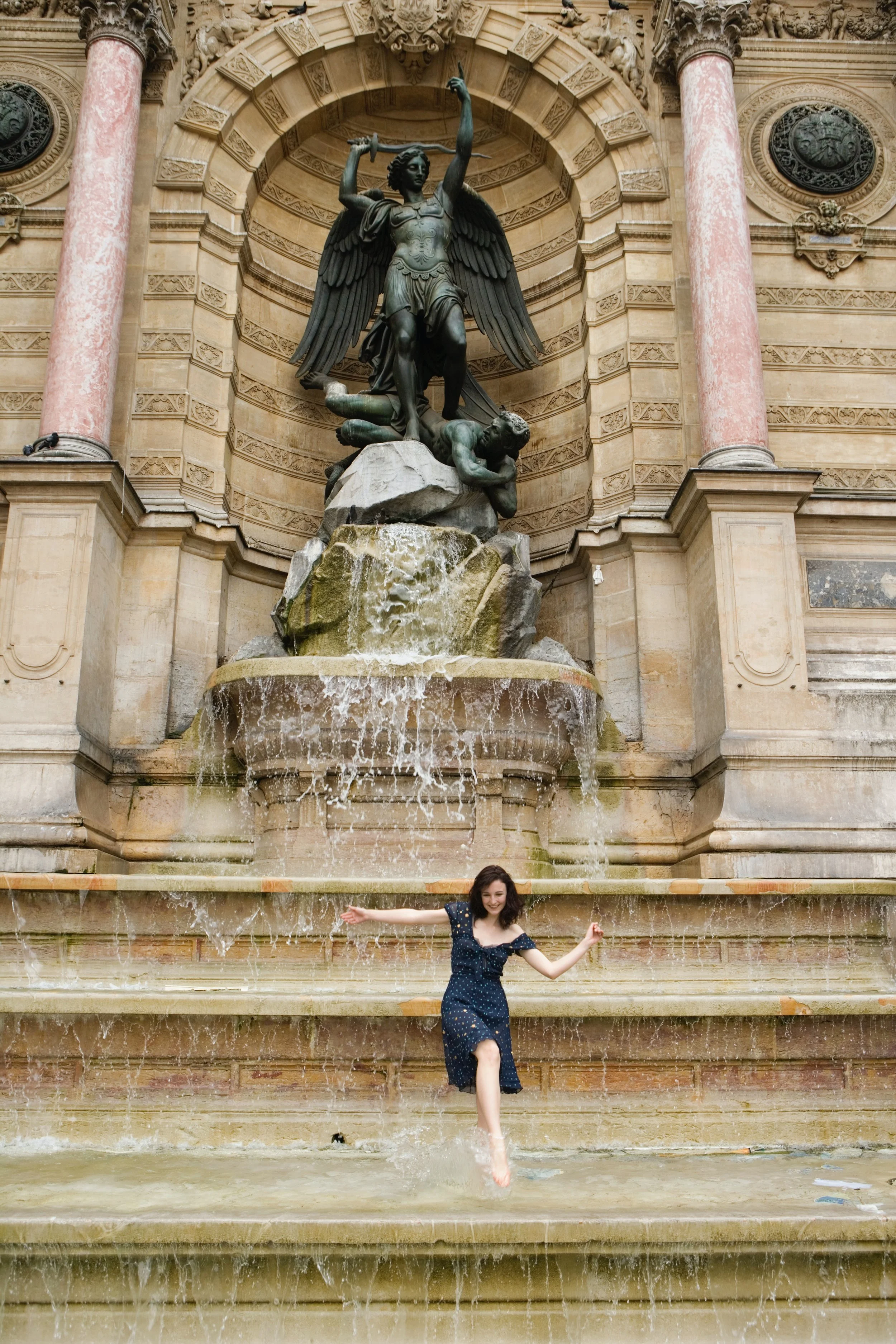 A young woman in a dark blue dress jumps and plays in front of a large ornate fountain with a sculpture of a winged figure holding a sword, surrounded by water features and intricate architectural details.