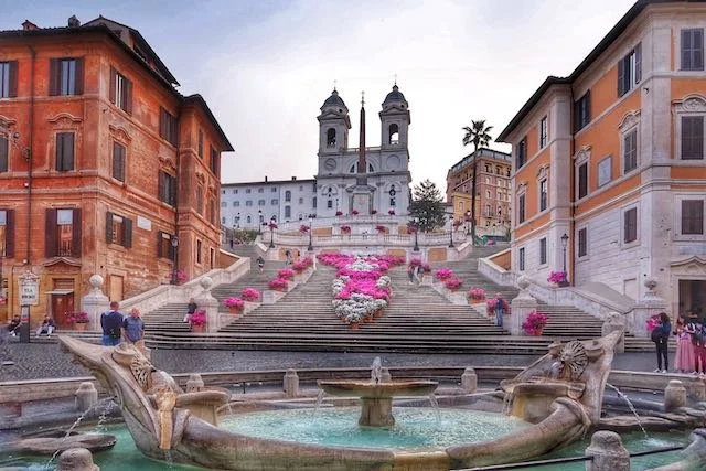 Colorful buildings surrounding the Spanish Steps with a fountain in the foreground, pink flowers decorating the stairs, and a church with twin bell towers at the top.