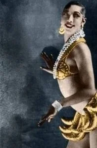 A woman dressed in vintage jewelry, gloves, and a dark-colored top, posing against a dark background.