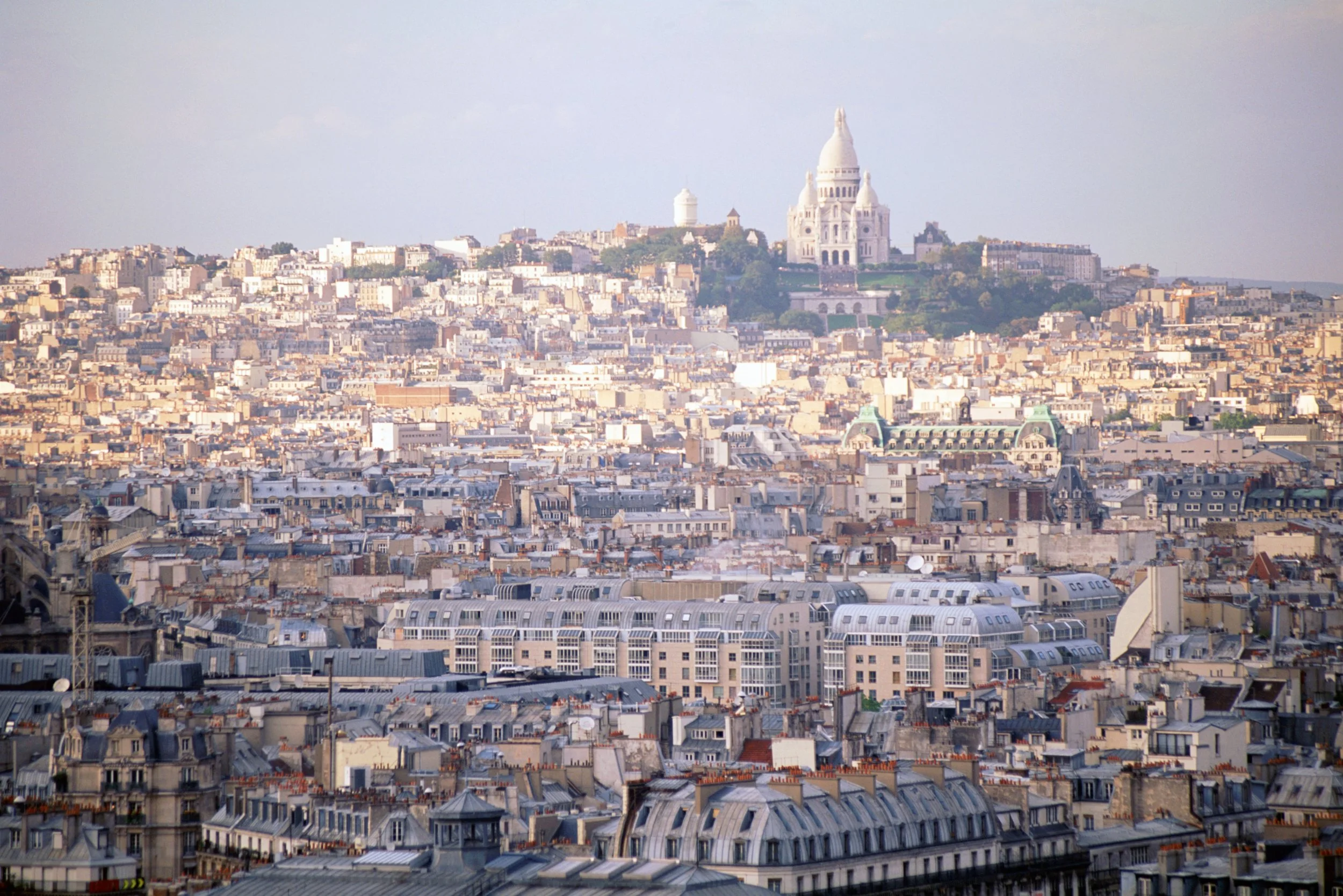 Panoramic view of Paris with the Basilica of Sacré-Cœur on Montmartre in the background and a densely built cityscape of rooftops and buildings in the foreground.