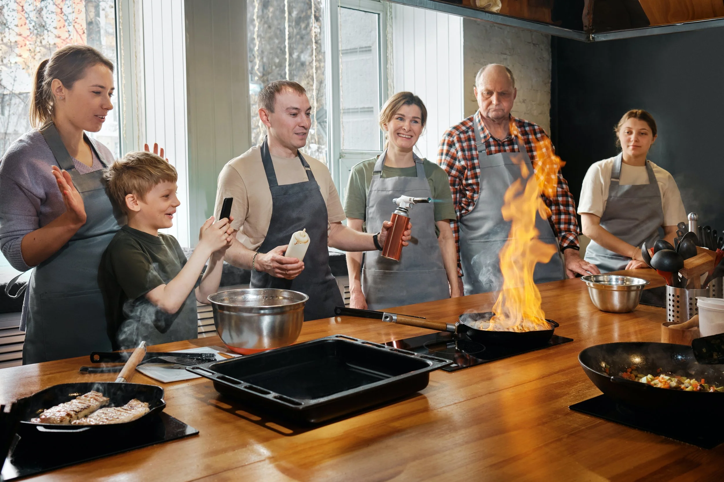 A group of people including children and adults in a kitchen, cooking on a stove with a large flame, while others watch and take pictures. They are wearing aprons and appear to be enjoying a cooking class or demonstration.
