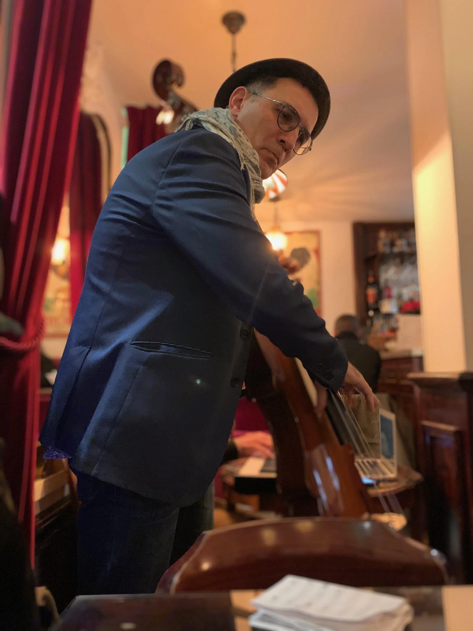 A man playing a double bass in a warmly lit restaurant or bar pub with red curtains and wooden furniture.