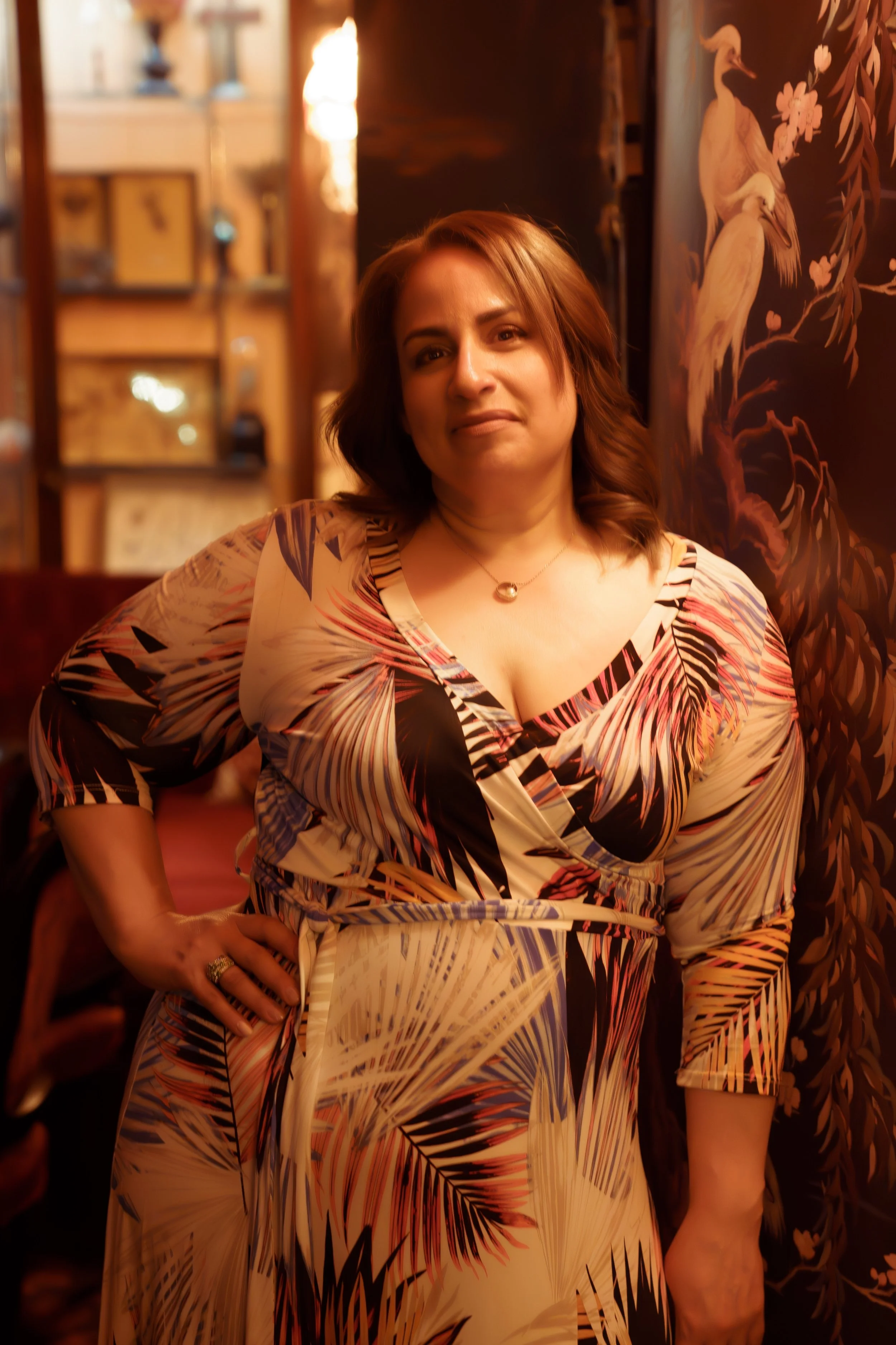 A woman with shoulder-length hair standing indoors in front of a decorative mural and shelves, wearing a patterned dress with tropical leaves and palm fronds.
