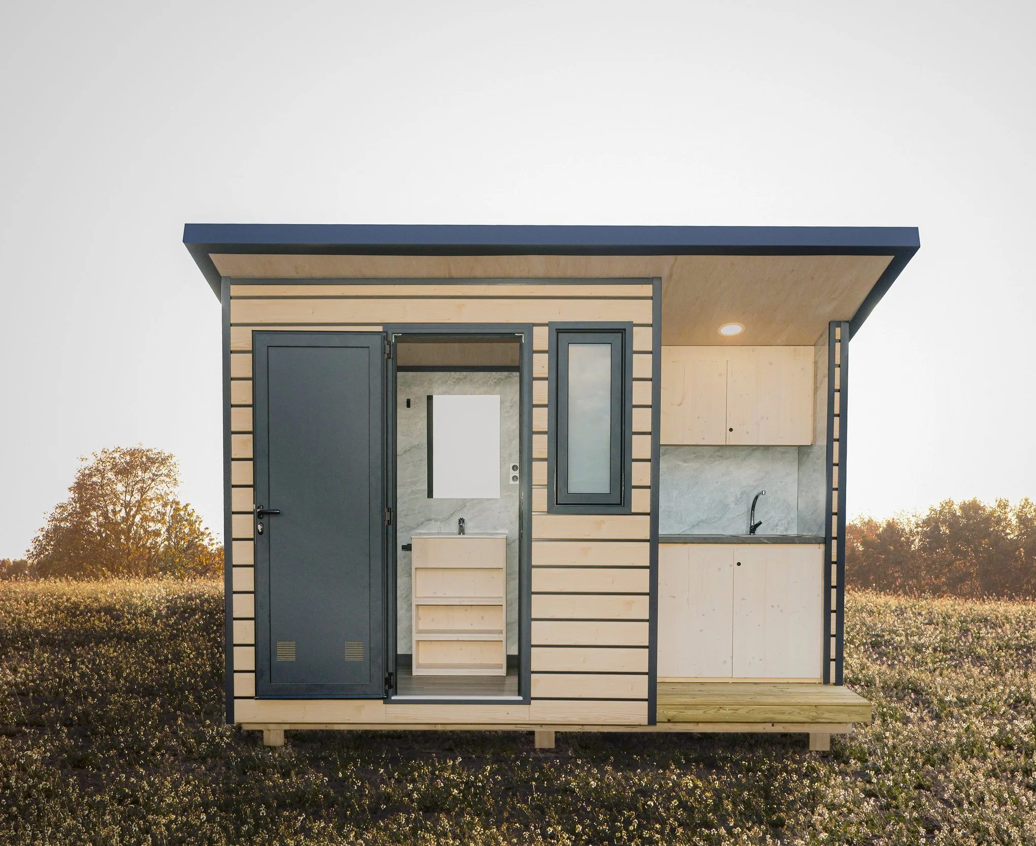 Pequeña cabaña Niuglam con paredes de madera, ventanas cuadradas y un pequeño lavabo, en un campo con árboles en el fondo.