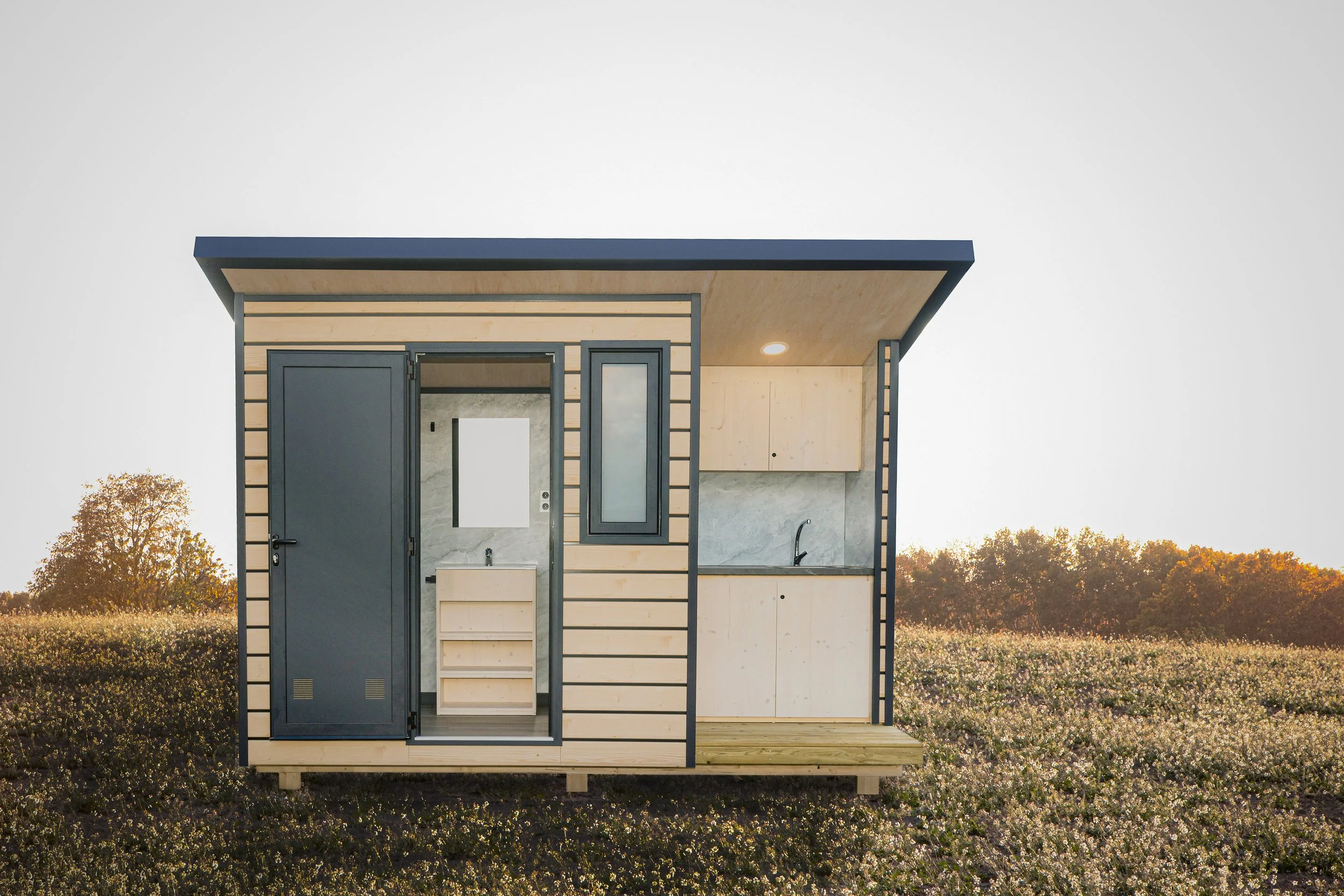 Modular Niuglam de madera moderno en un campo con árboles al fondo, con paredes de madera, puerta metálica, ventana y cocina en el interior