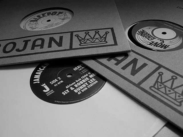 Three vinyl Records in Trojan sleeves on a table, featuring the Trojan logo in muted grey tones, representing the foundations of reggae and Jamaican sound system culture.