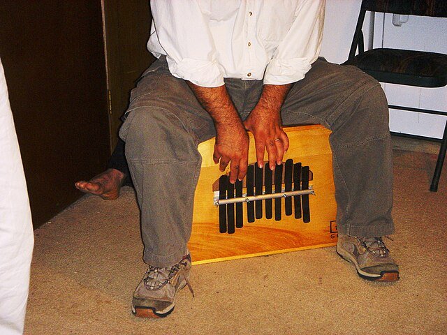Man sitting on a Rumba Box percussion instrament, playing it, shown from the waist down.