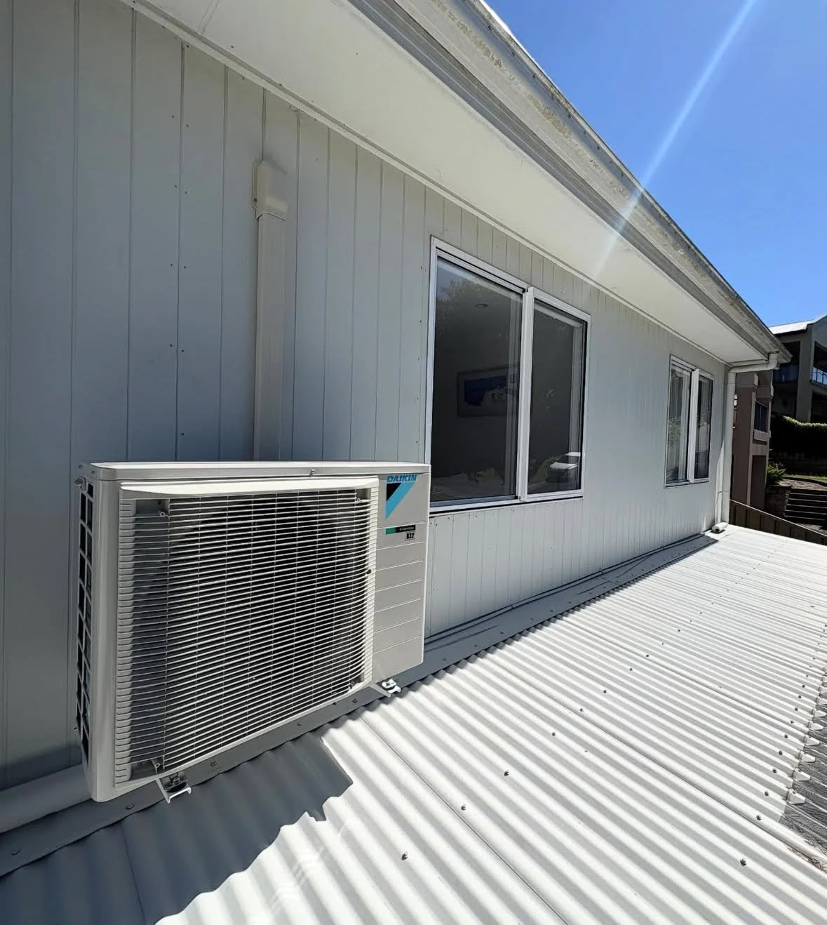 Exterior view of a white building with a Daikin air conditioning unit mounted on the corrugated metal roof, two large windows, and clear blue sky.