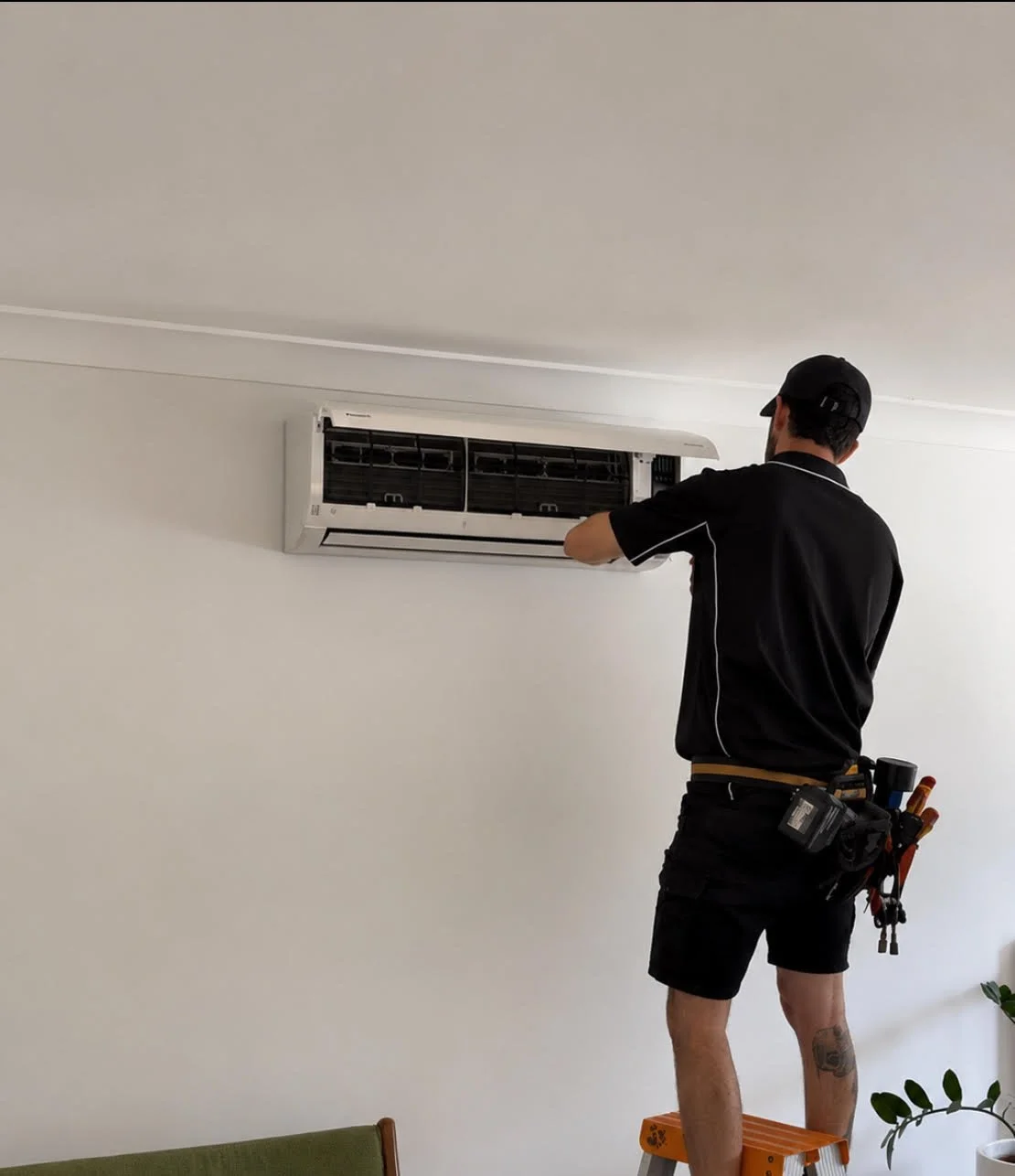 A technician repairing a wall-mounted air conditioning unit in a room with white walls, standing on a small orange step ladder, with tools attached to his belt.