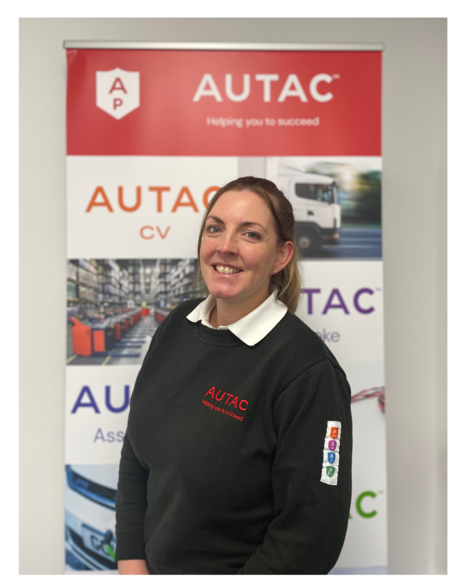 Image of Sarah Bates, Finance Manager stood in front of Autac banner wearing black jumper with Autac logo. 
