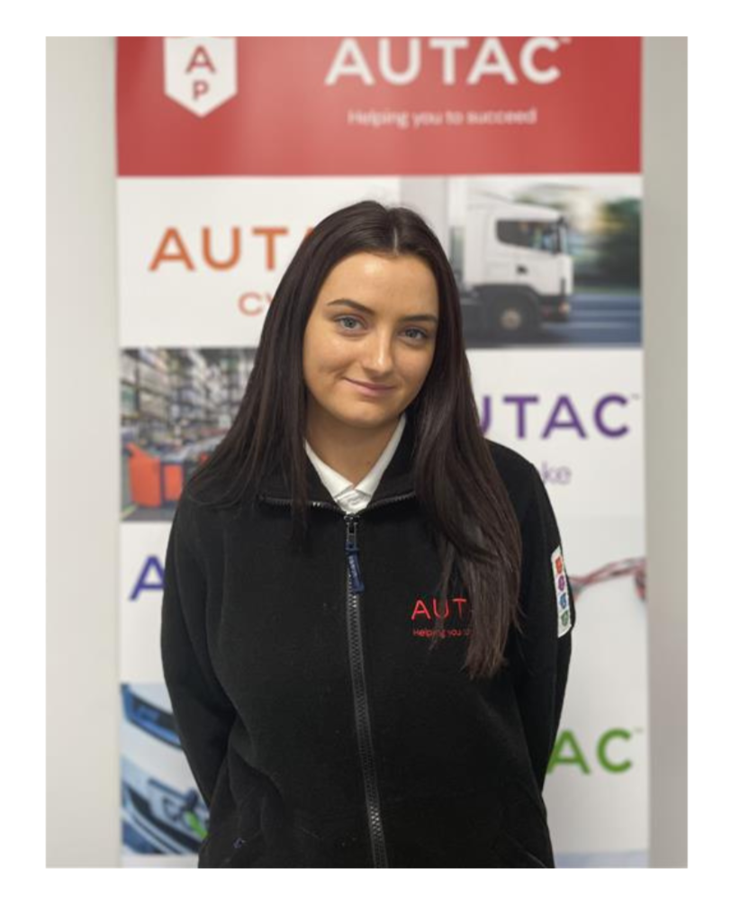 Image of Darcey O'Hara, Office Administrator stood in front of Autac banner wearing black fleece with Autac logo. 