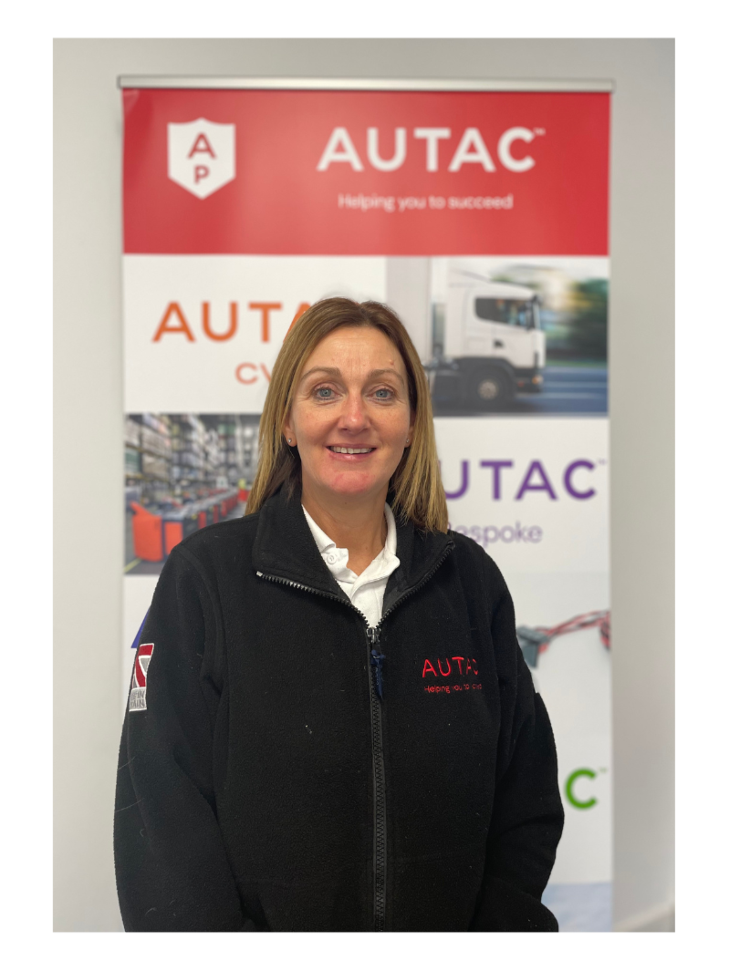 Image of Sallie Lowe, Finance Director,  stood in front of Autac banner wearing black fleece with Autac logo. 