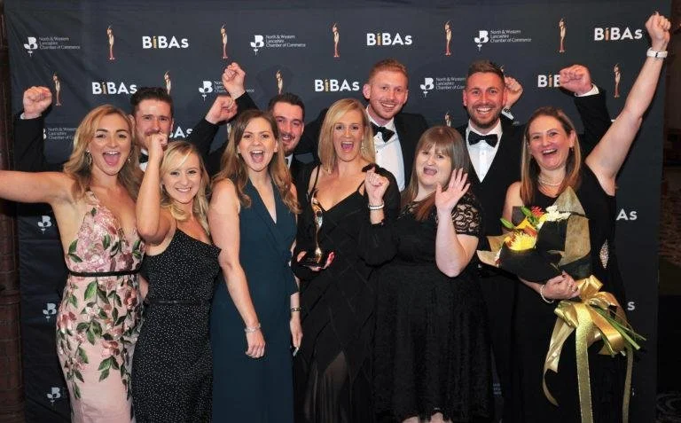 A group of people joyfully celebrating at an awards event, standing in front of a "BIBAs" backdrop, with one person holding a bouquet.