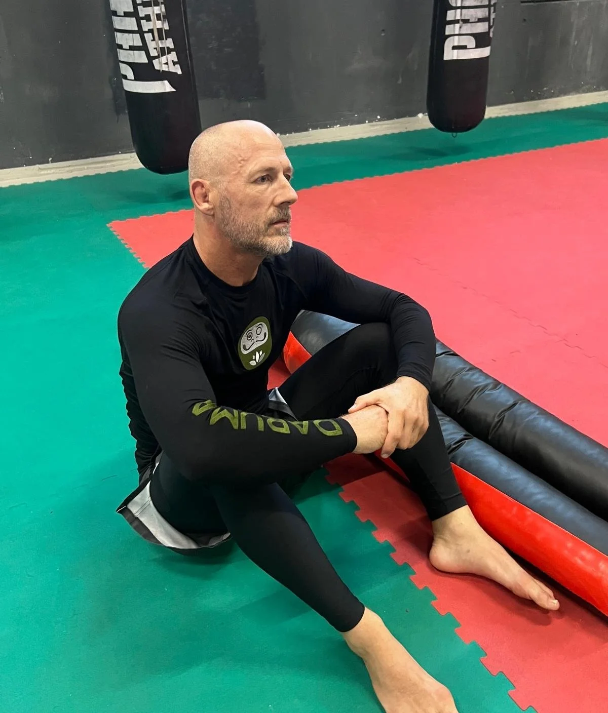 A man with a bald head and beard sitting on the mat in a gym, with punching bags hanging behind him, wearing a black long-sleeve shirt and black leggings.