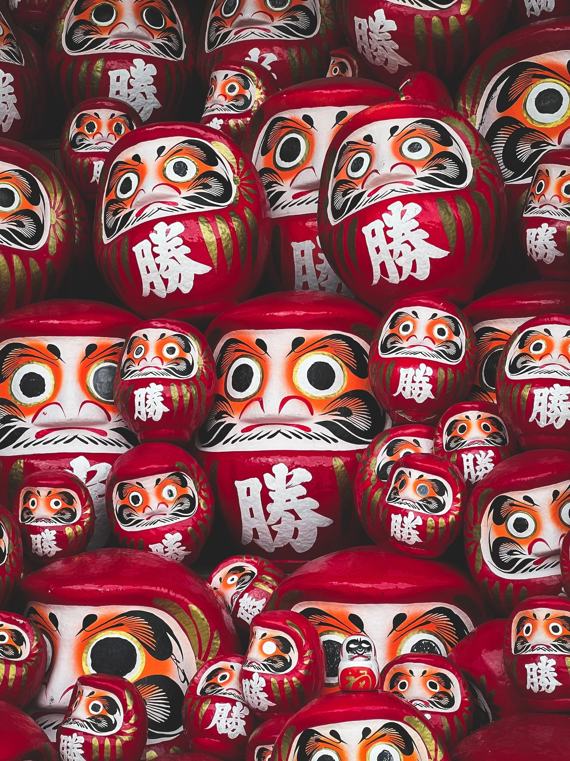 A collection of red Daruma dolls with white faces, large eyes, and black, orange, and gold painted features, stacked together.