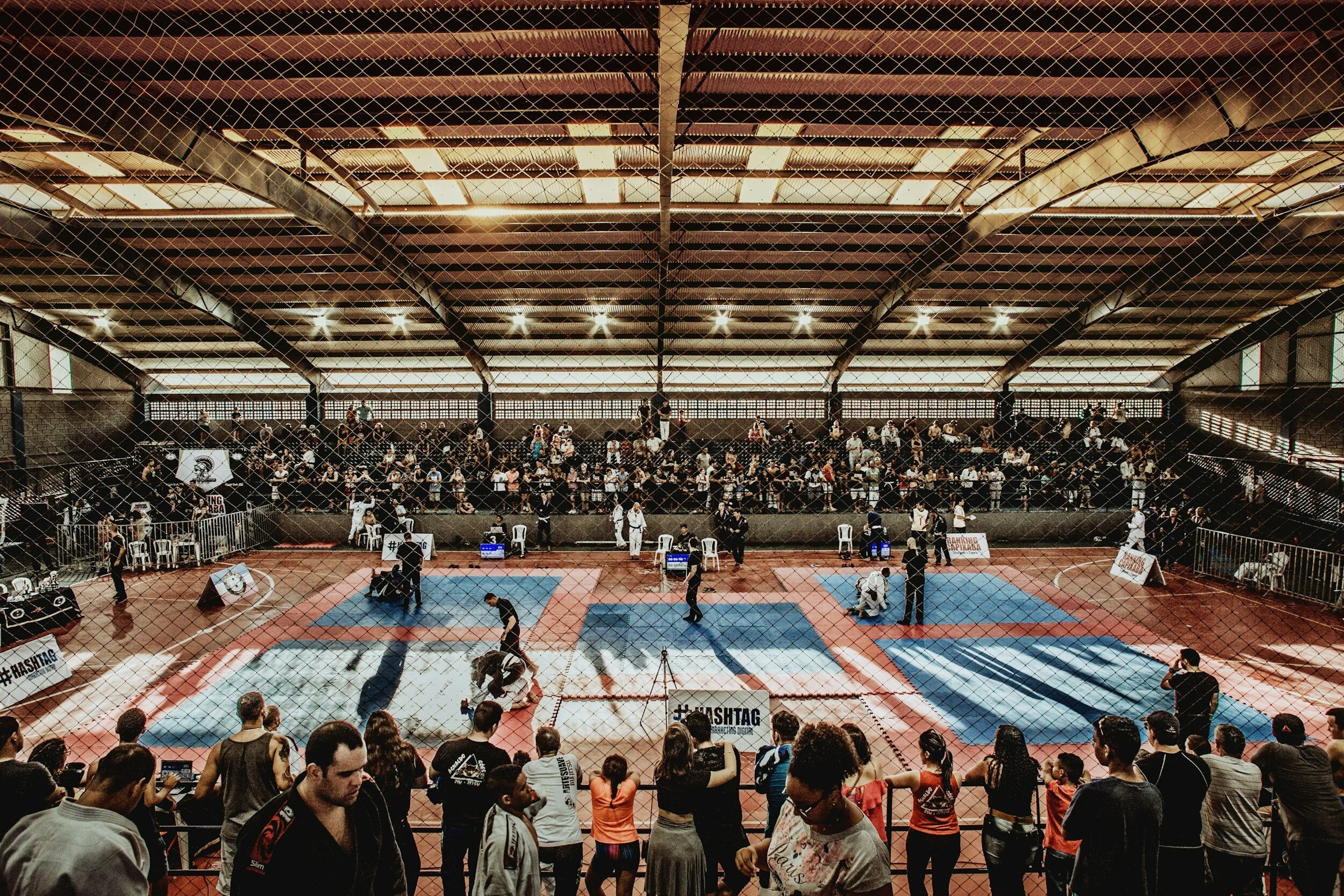 An indoor martial arts or combat sports tournament with competitors fighting in several matches, with spectators watching from the stands behind a protective net.