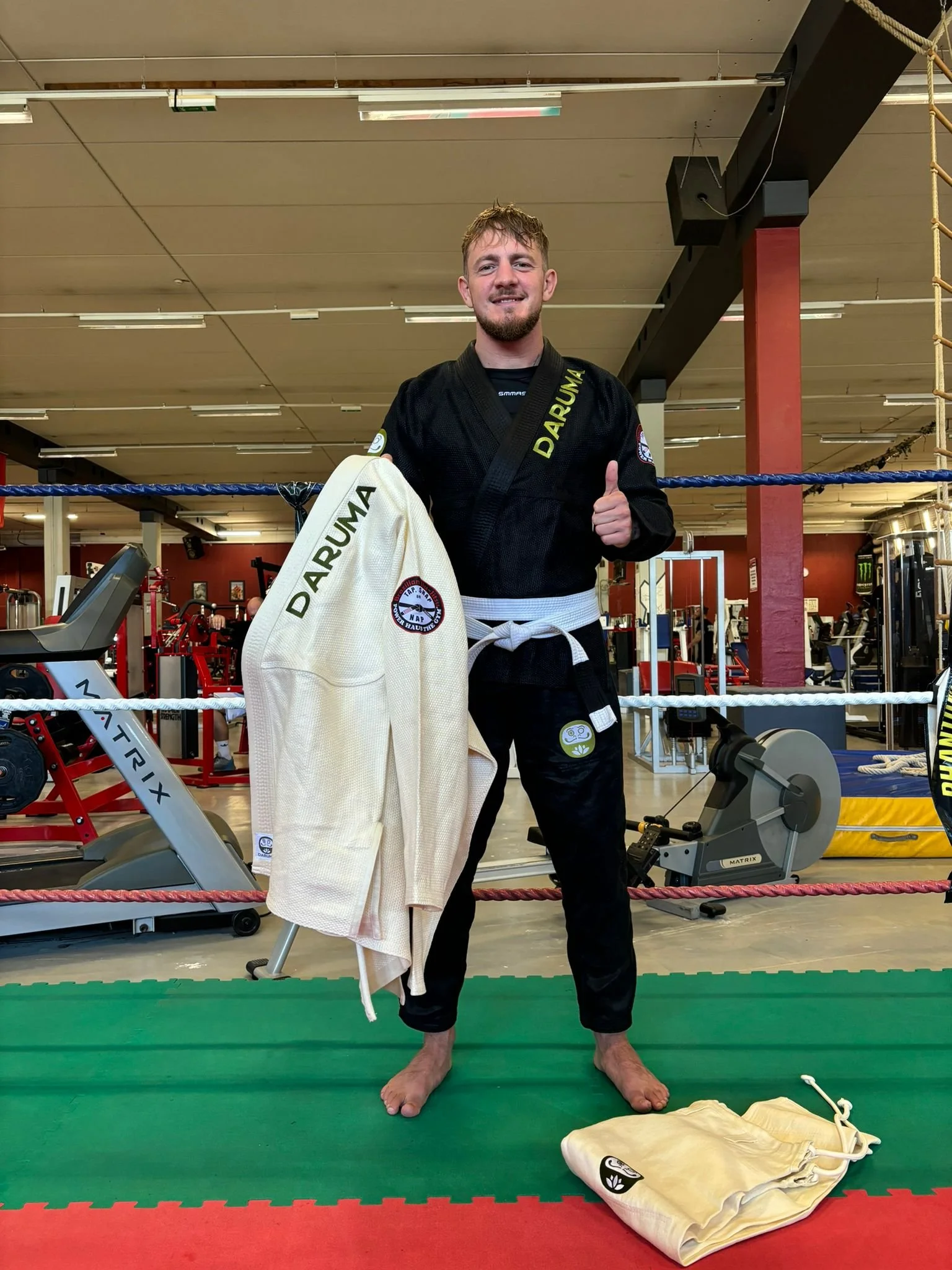 Man wearing a black jiu-jitsu gi, holding a white belt and a gis jacket with 'DARUMA' written on it, standing inside a gym with various exercise equipment.
