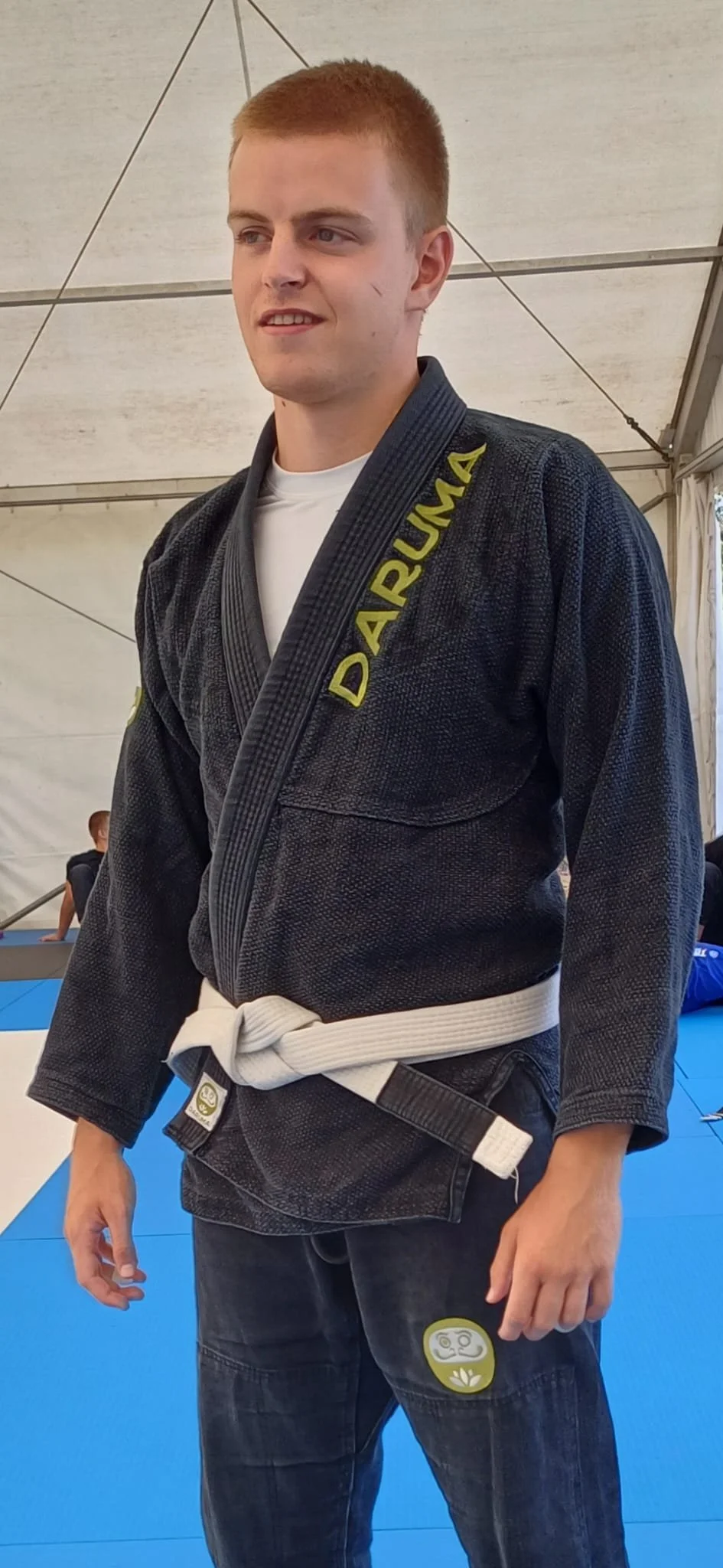 Male Brazilian Jiu-Jitsu competitor wearing a black gi with yellow 'DARUMA' lettering, white belt, and black jeans, standing on blue mat in tented area.