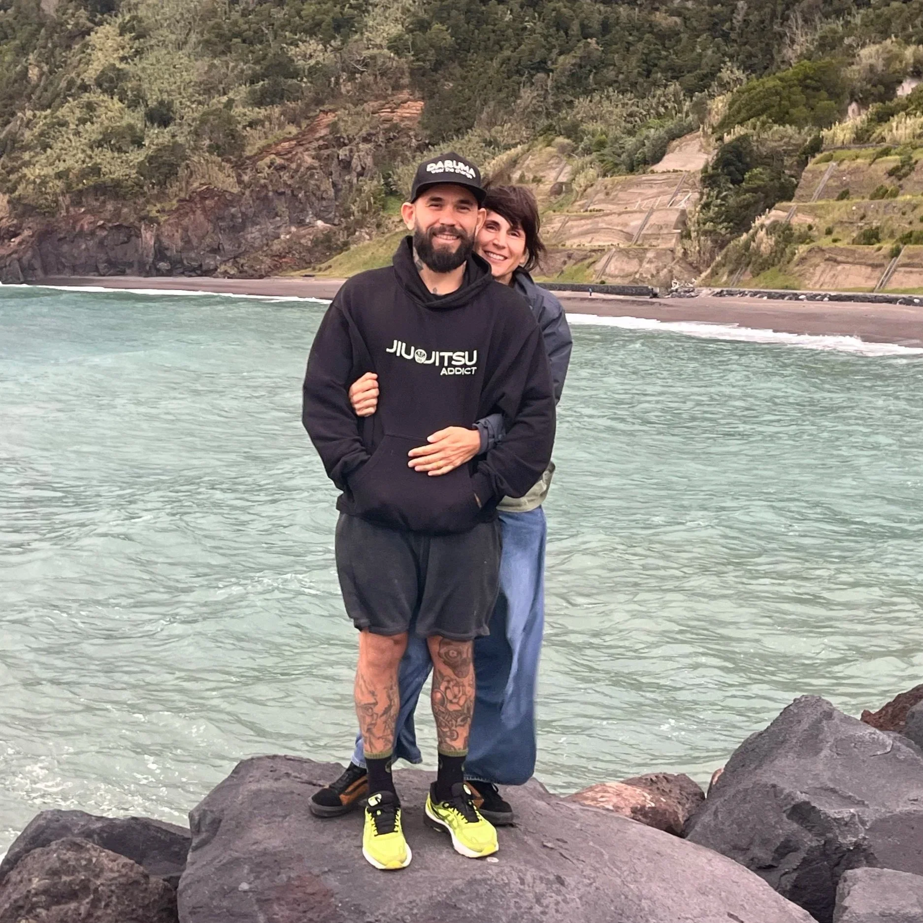 A man and woman standing on rocks by the ocean, hugging each other and smiling. The background shows water, a beach, and hillside with trees and terraced landscape.
