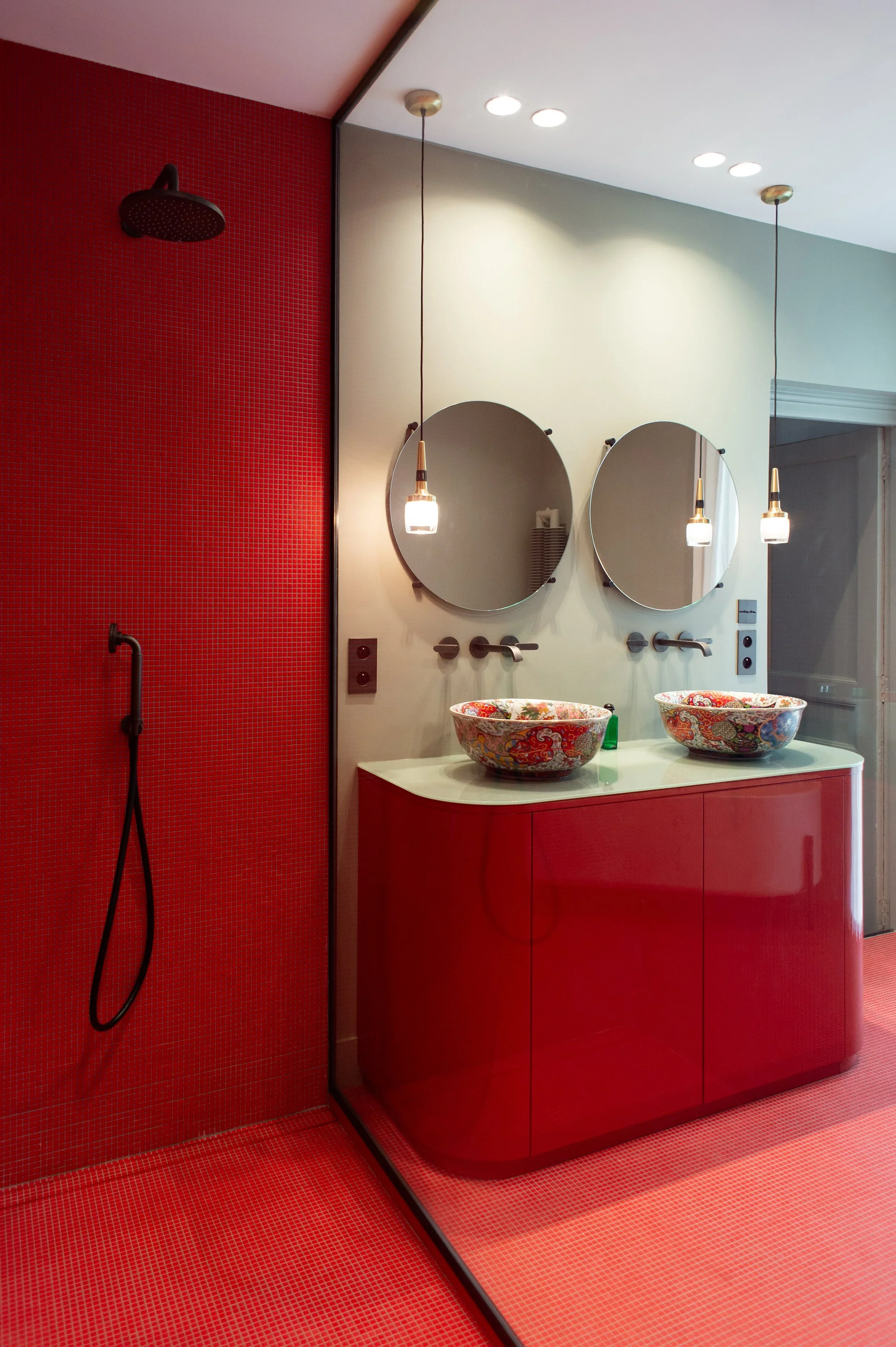 Salle de bain avec un mur rouge, deux vasques colorées sur un meuble rouge, deux miroirs ronds, deux suspensions lumineuses, une douche à l'italienne avec un panneau de verre.