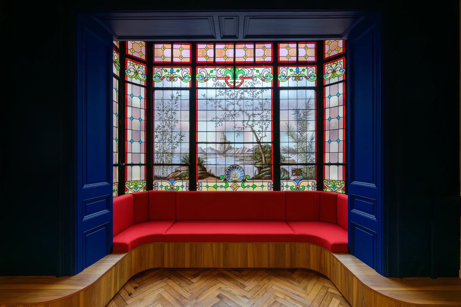 Une fenêtre en vitrail coloré représentant un paysage marin avec des palmiers, un rocher, une tour et des oiseaux, encadrée par des volets bleus, avec un siège rouge courbé en dessous et un sol en parquet en bois.