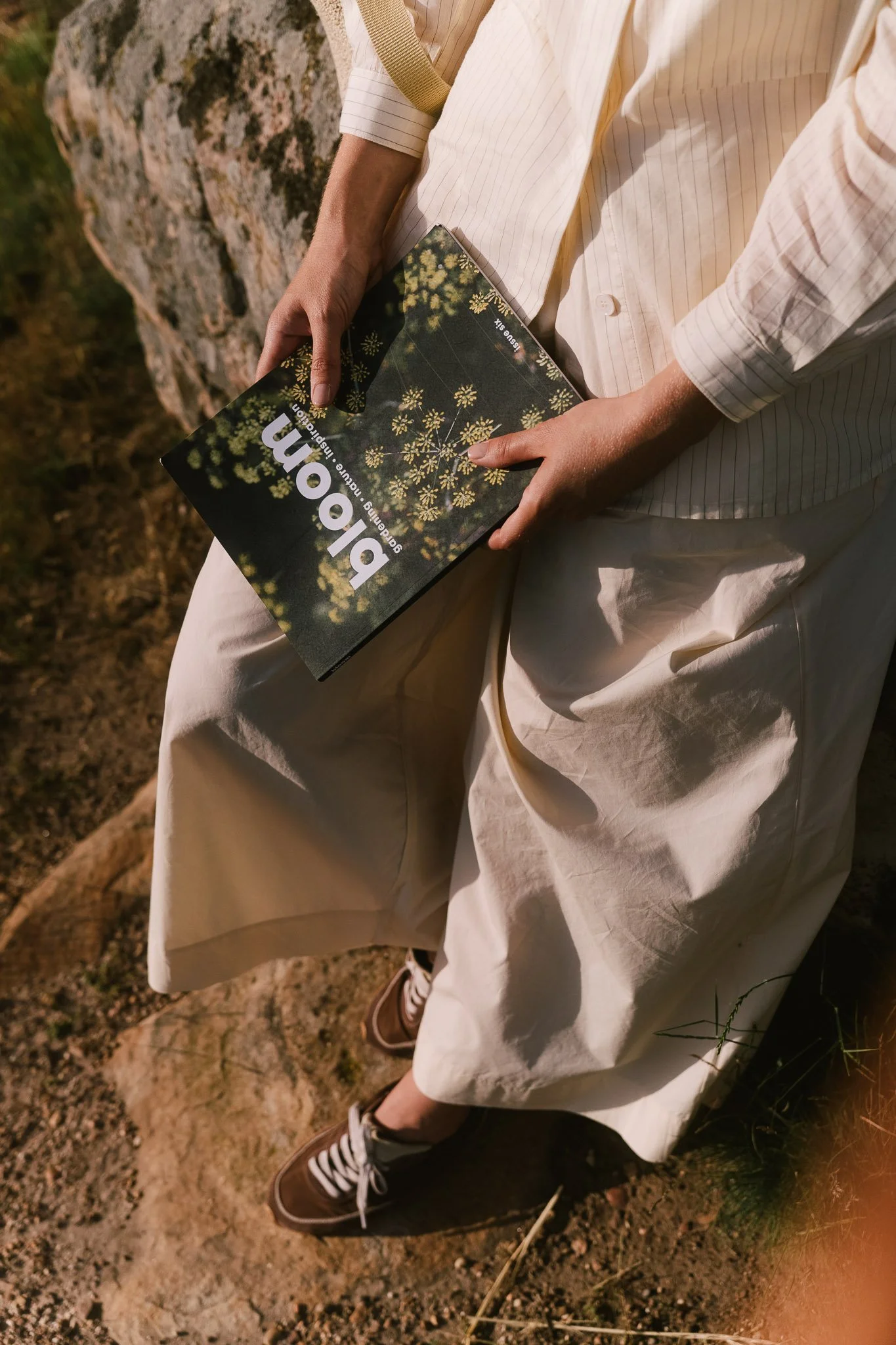 Krémszínű nadrágot viselő alak kőlépcsőn ül és egy „bloom" című magazint tart, meleg napfényben