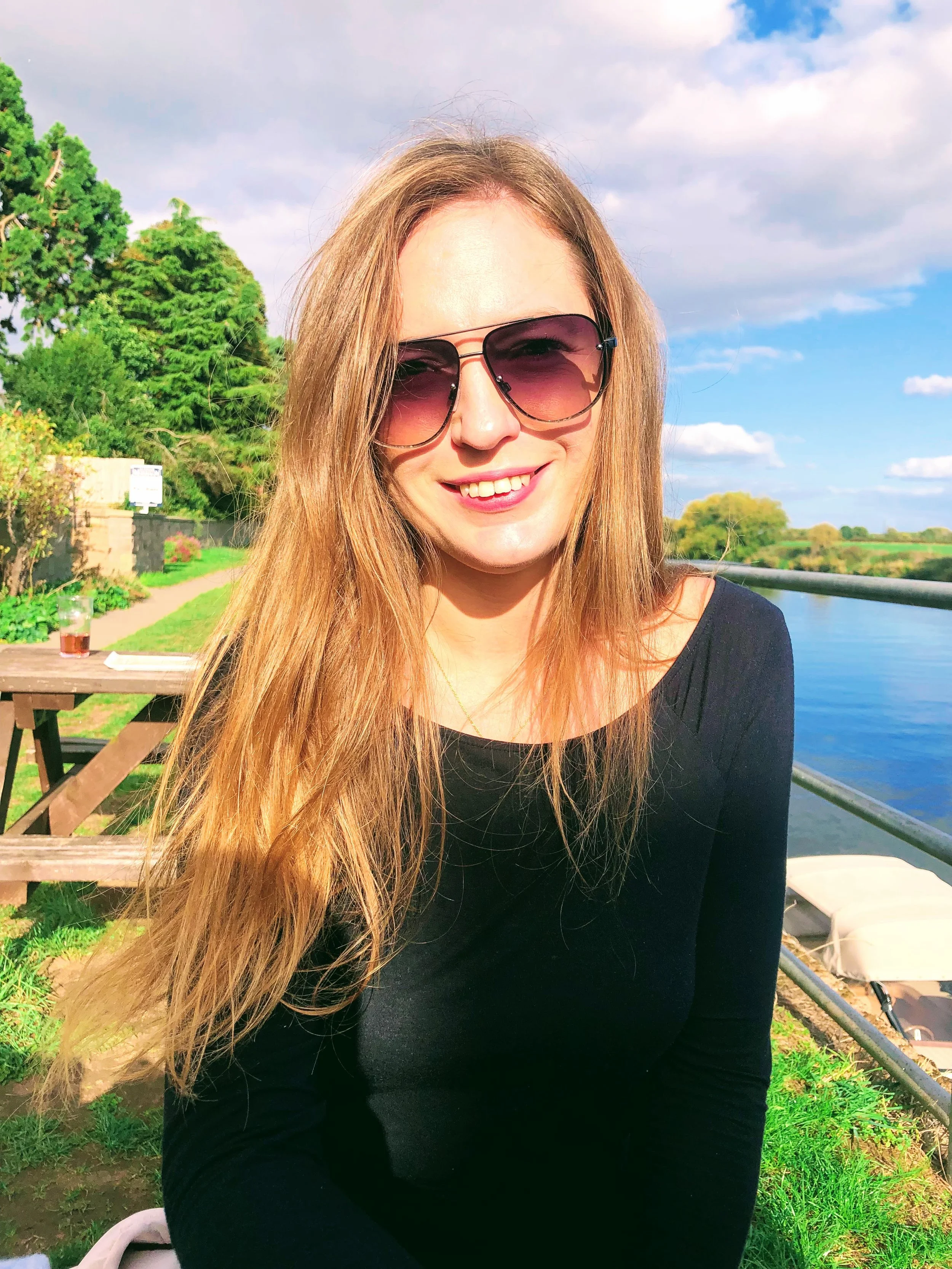 Une femme souriante portant des lunettes de soleil, assise à l'extérieur près d'une rivière avec un ciel partiellement nuageux, entourée de verdure.