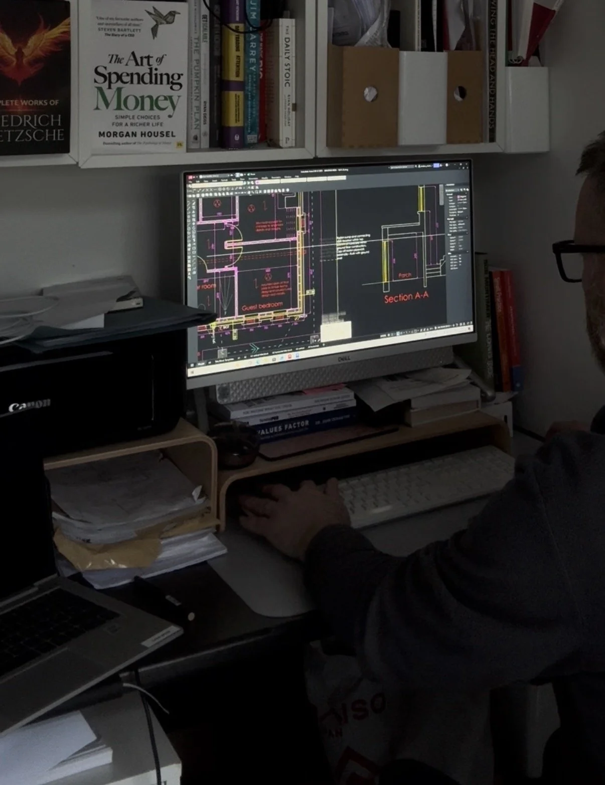 Person working on architectural drawings on a computer screen, with books and office supplies on the desk and shelves.