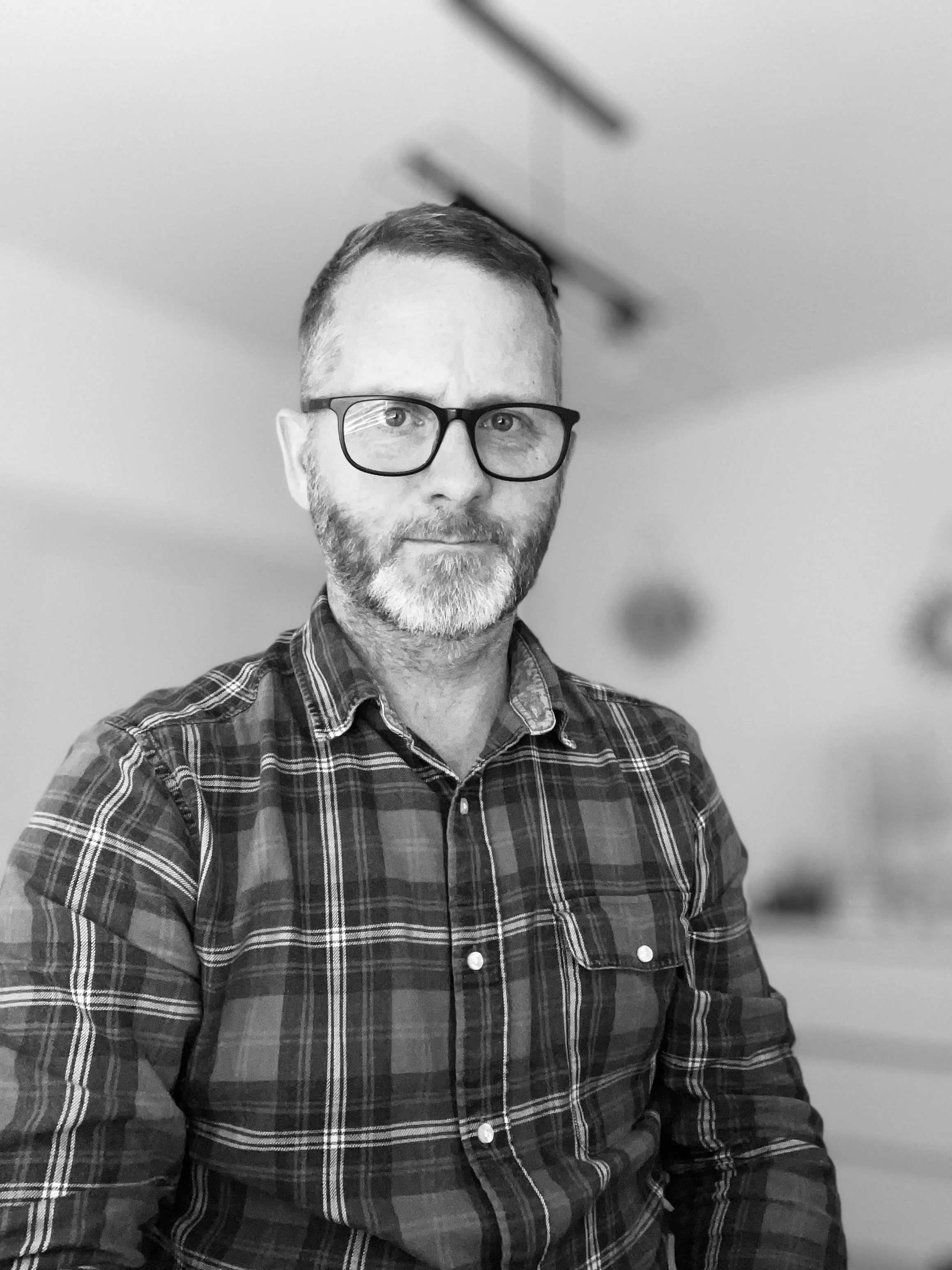 A black and white photo of a middle-aged man with glasses and a beard, wearing a plaid shirt, sitting indoors with a plain wall and ceiling visible in the background.