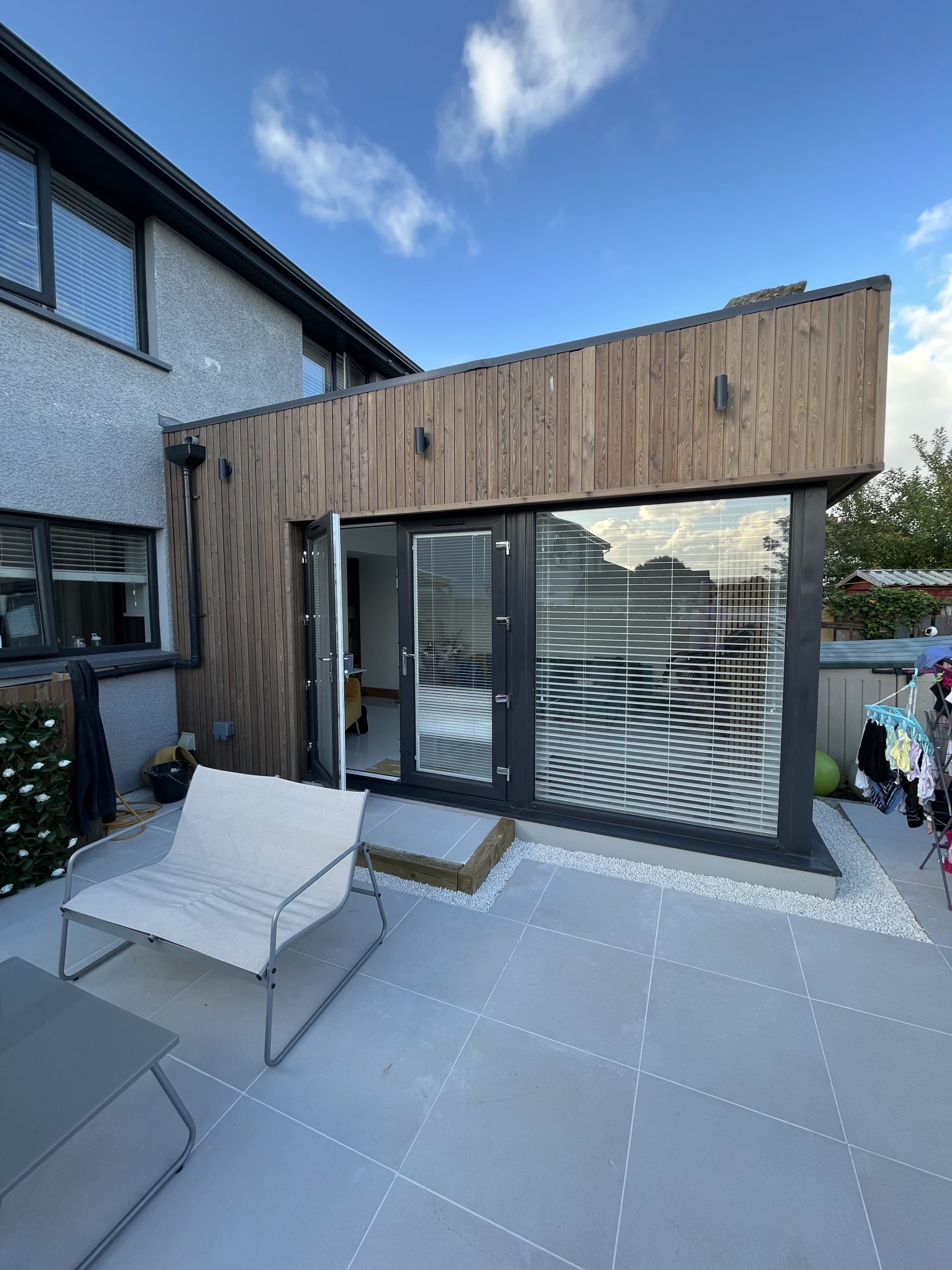 A modern residential backyard patio with a lounge chair, a small table, a garden bed, and an extension of the house with large windows and a glass door. The house features a two-story design with a gray and wood exterior, and the sky is blue with some clouds.