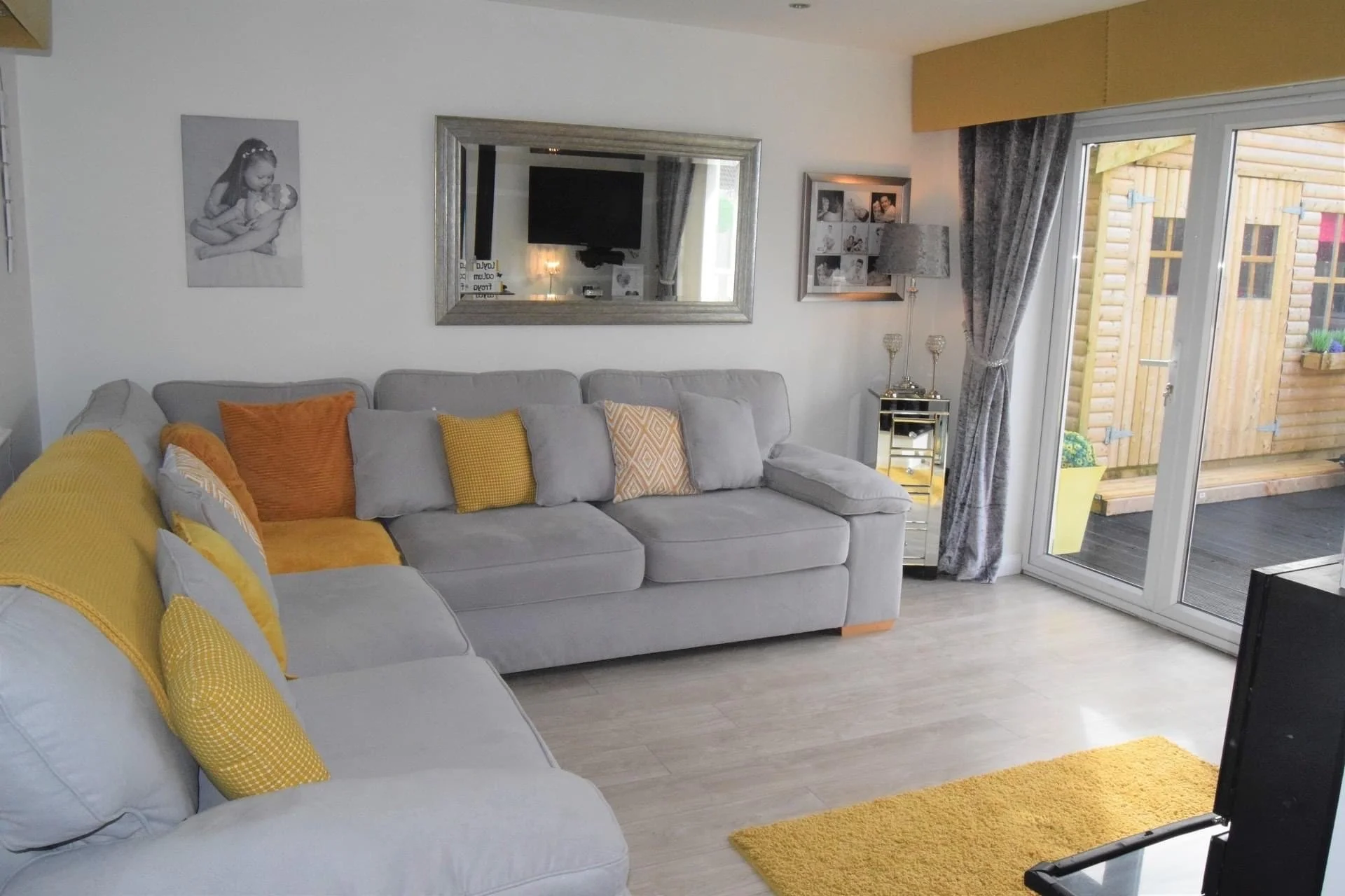 Living room with light gray sofa adorned with yellow and patterned throw pillows, yellow blanket, large mirror on white wall reflecting a TV and window, framed collage of black and white photos, silver side table with candles and a lamp, sliding glass door leading outside to a wooden deck with potted plants, gray curtains, light-colored wooden flooring, yellow rug.