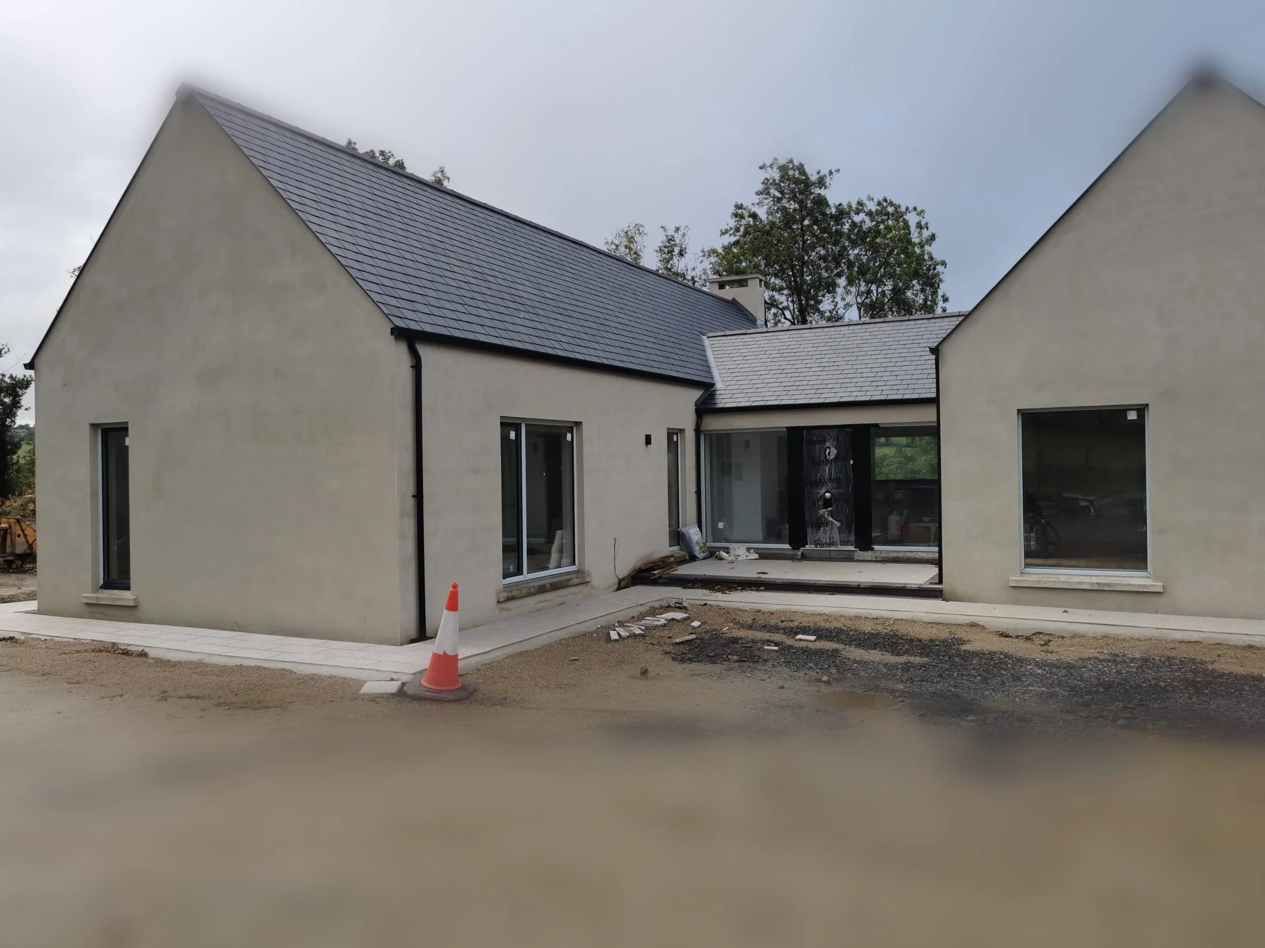 A house under construction with finished exterior walls, a steep roof with dark tiles, and large sliding glass doors. There is a construction cone on the ground in front of the house, and the area around it is still being developed.