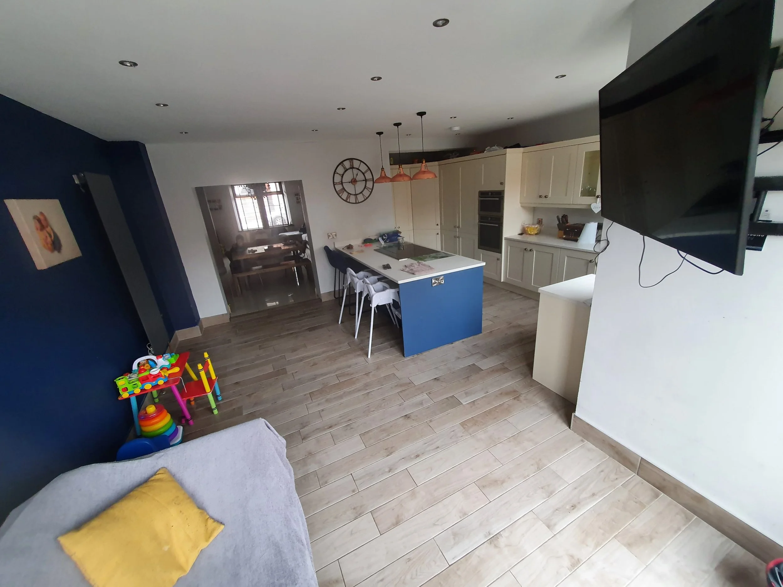 View of a modern kitchen and dining area with a blue accent wall, a children's toy on the floor, white cabinets, and a large flat-screen TV mounted on the wall.