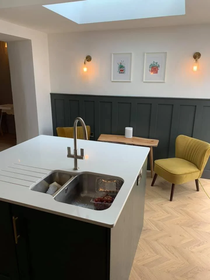 Modern kitchen island with stainless steel sink, faucet, and a few grapes inside. In the background, there is a small wooden table with a white speaker, and two yellow chairs. The wall behind has dark paneling with two framed artprints and wall-mounted light fixtures.