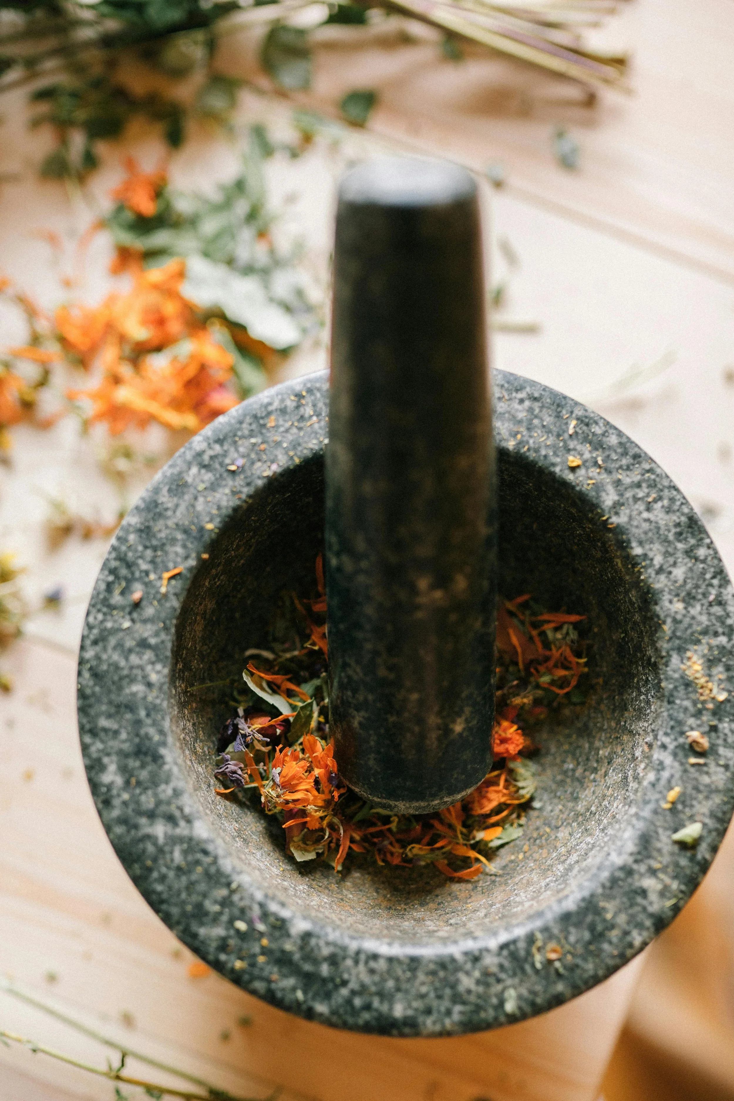 Close-up of a mortar and pestle filled with crushed dried flowers, with scattered flower petals around it on a wooden surface.
