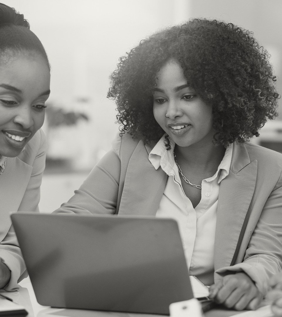 Two women in business attire looking at a laptop together in an office setting.