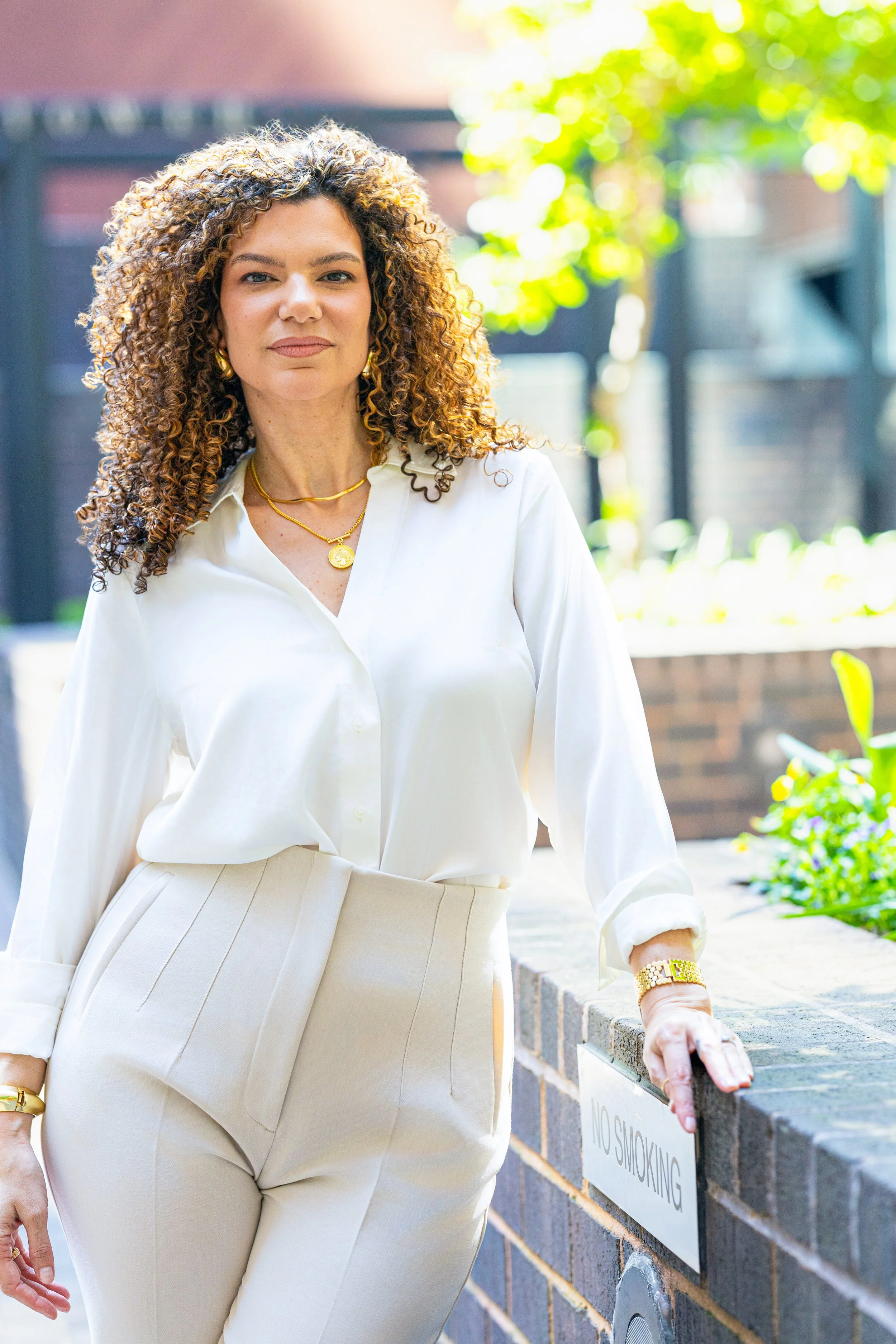 A woman with curly hair wearing a white blouse and beige pants, standing outdoors near a brick wall with a 'No Smoking' sign, on a sunny day with blurred greenery in the background.